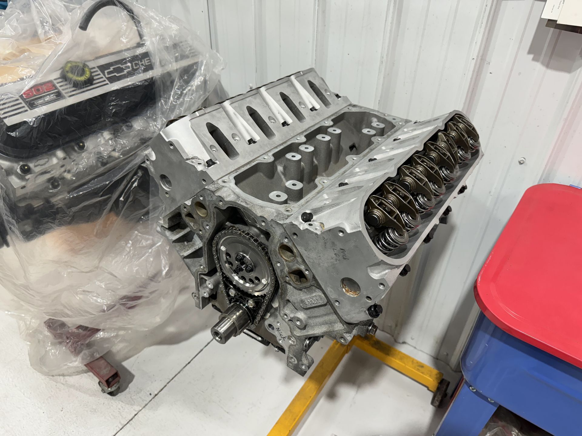 Car engine block on a workshop floor beside plastic-wrapped parts and a red-and-blue tool cart