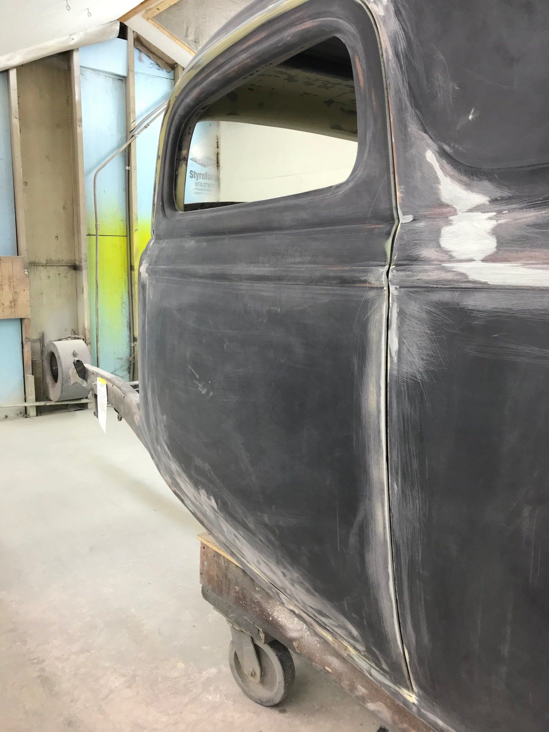 Black unfinished vehicle body on wheels in a workshop, with a window opening and dusty floor.