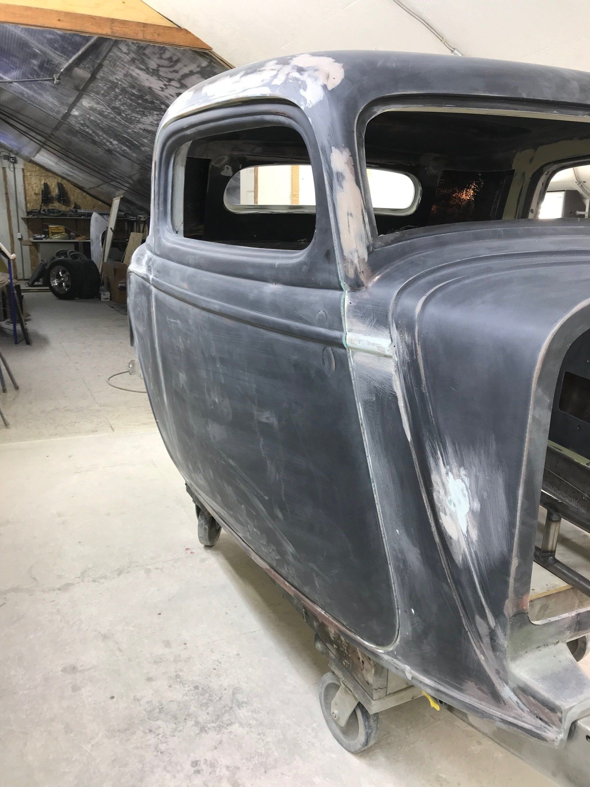 Rusty black vintage car body in a workshop, missing windows and front parts, on wheels.