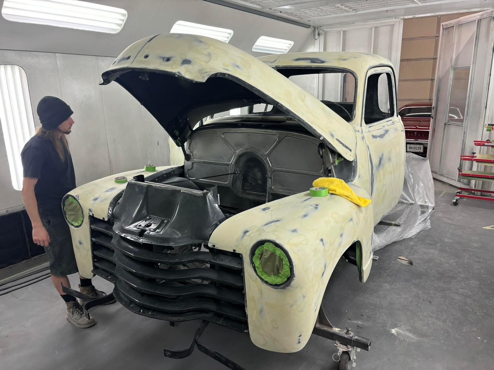 Classic pickup truck in a spray booth, hood open and partially restored, with a worker beside it