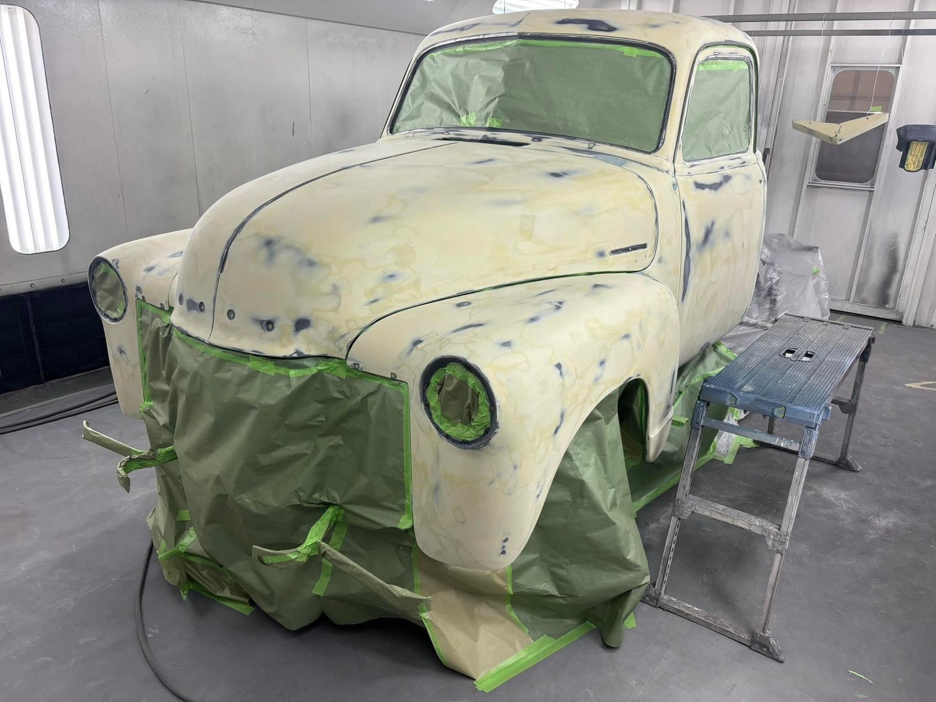 Old car body in a paint booth, masked with green plastic and beige primer, with a step ladder nearby