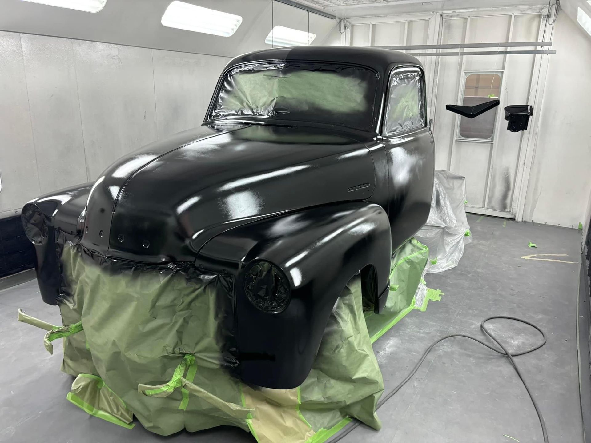 Black vintage pickup truck being painted in a spray booth, masked with green tape and paper.