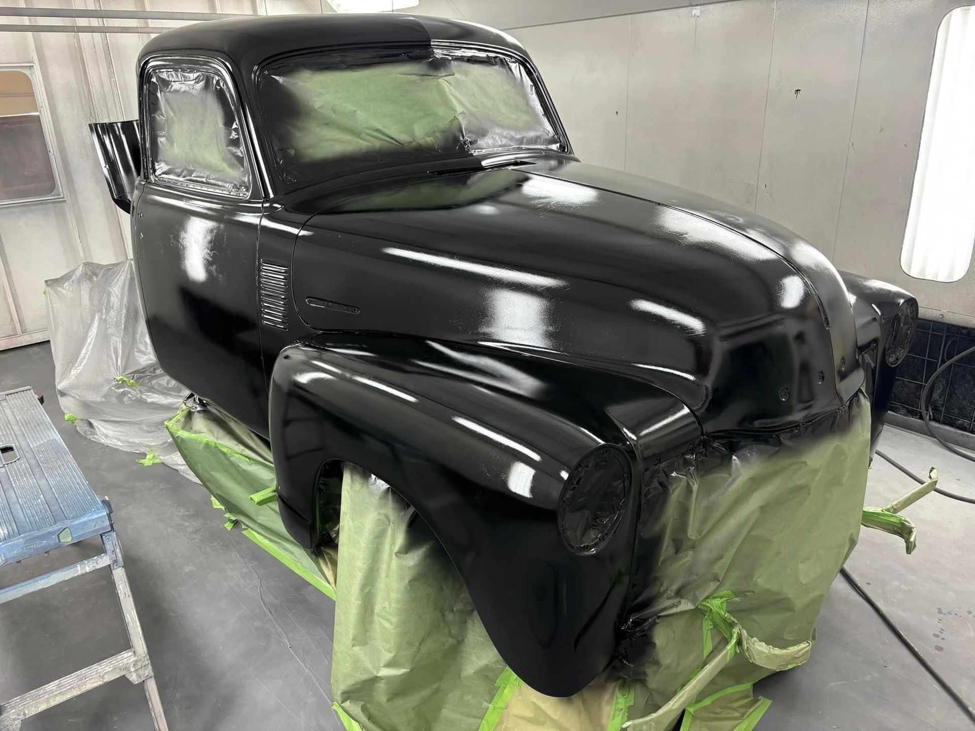 Glossy black vintage pickup truck cab and hood masked for painting in a spray booth