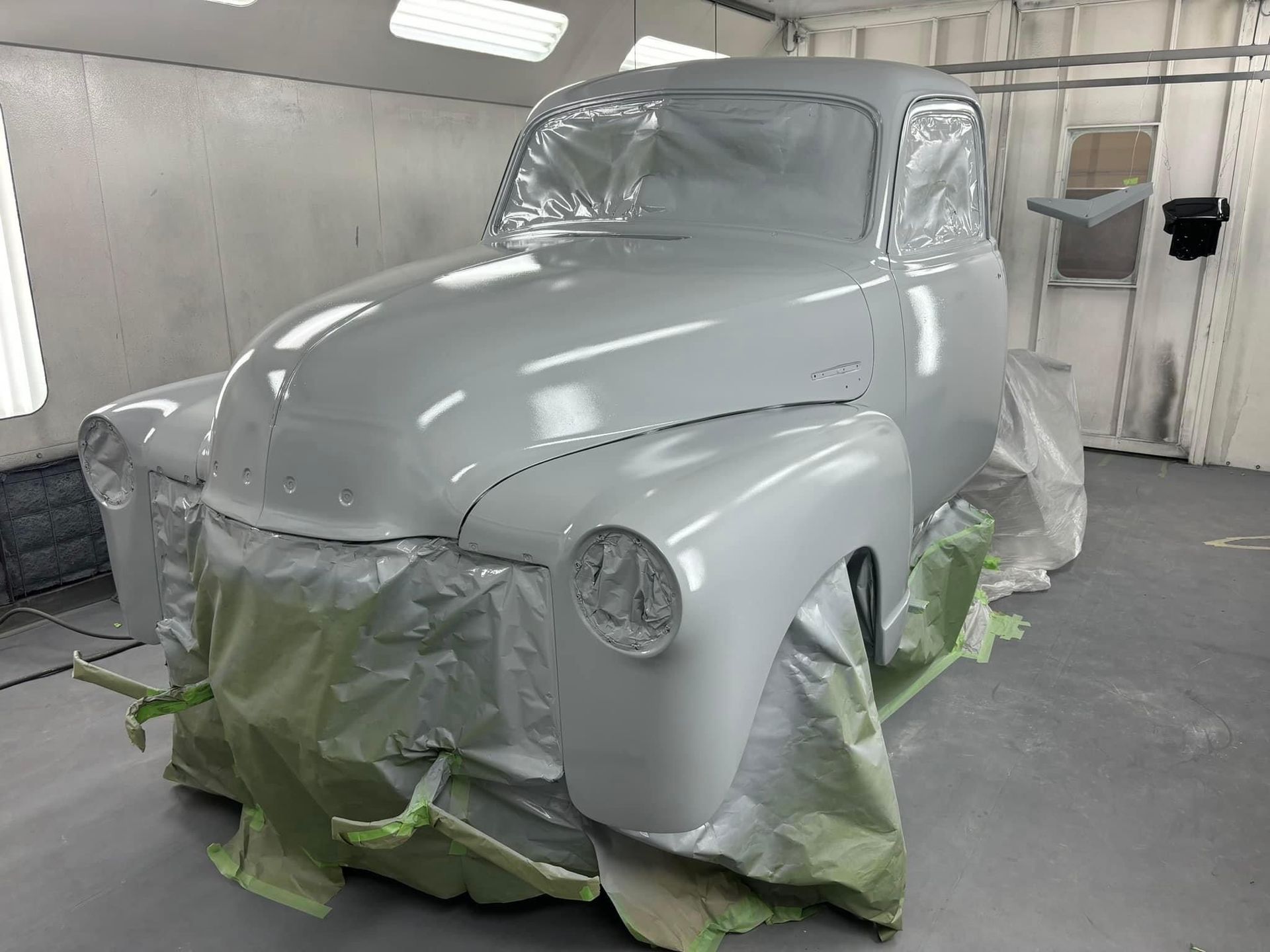 Gray classic car body in a paint booth, masked with green tape and paper, awaiting paint