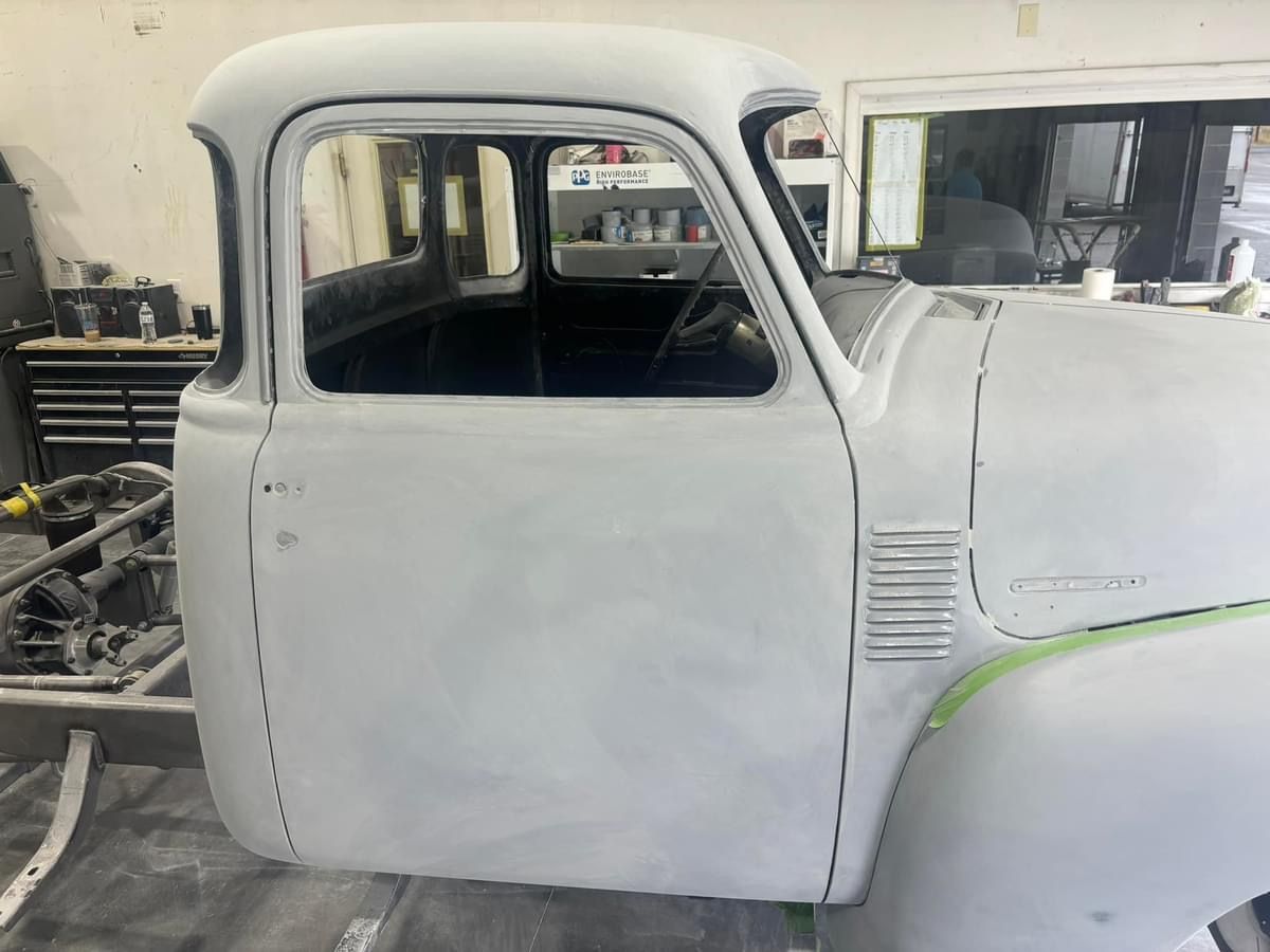 Primed vintage pickup cab in a workshop, white body with the hood and front fender partially visible.