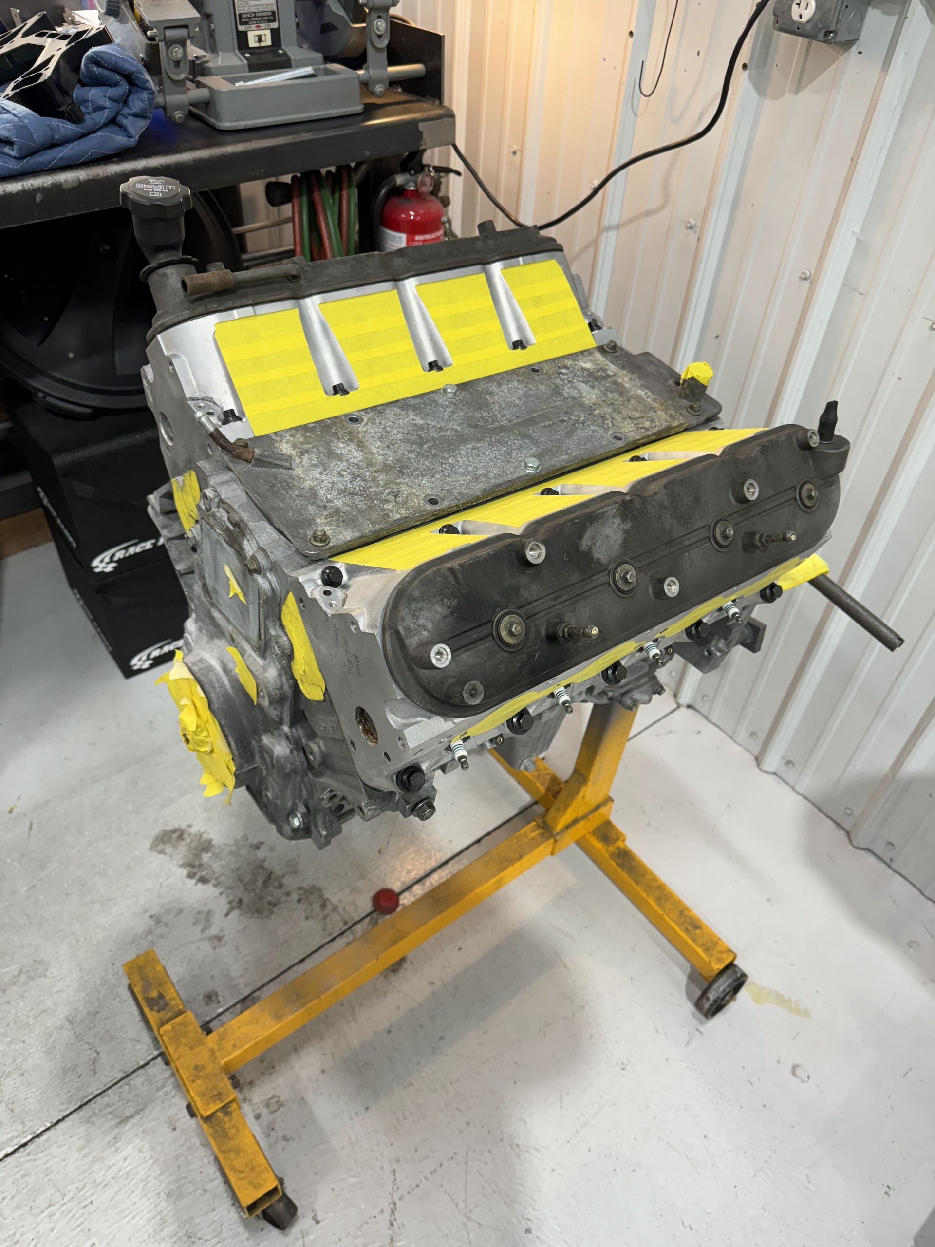 Yellow engine block on a stand in a workshop, wrapped in gray insulation with black mounting plate.