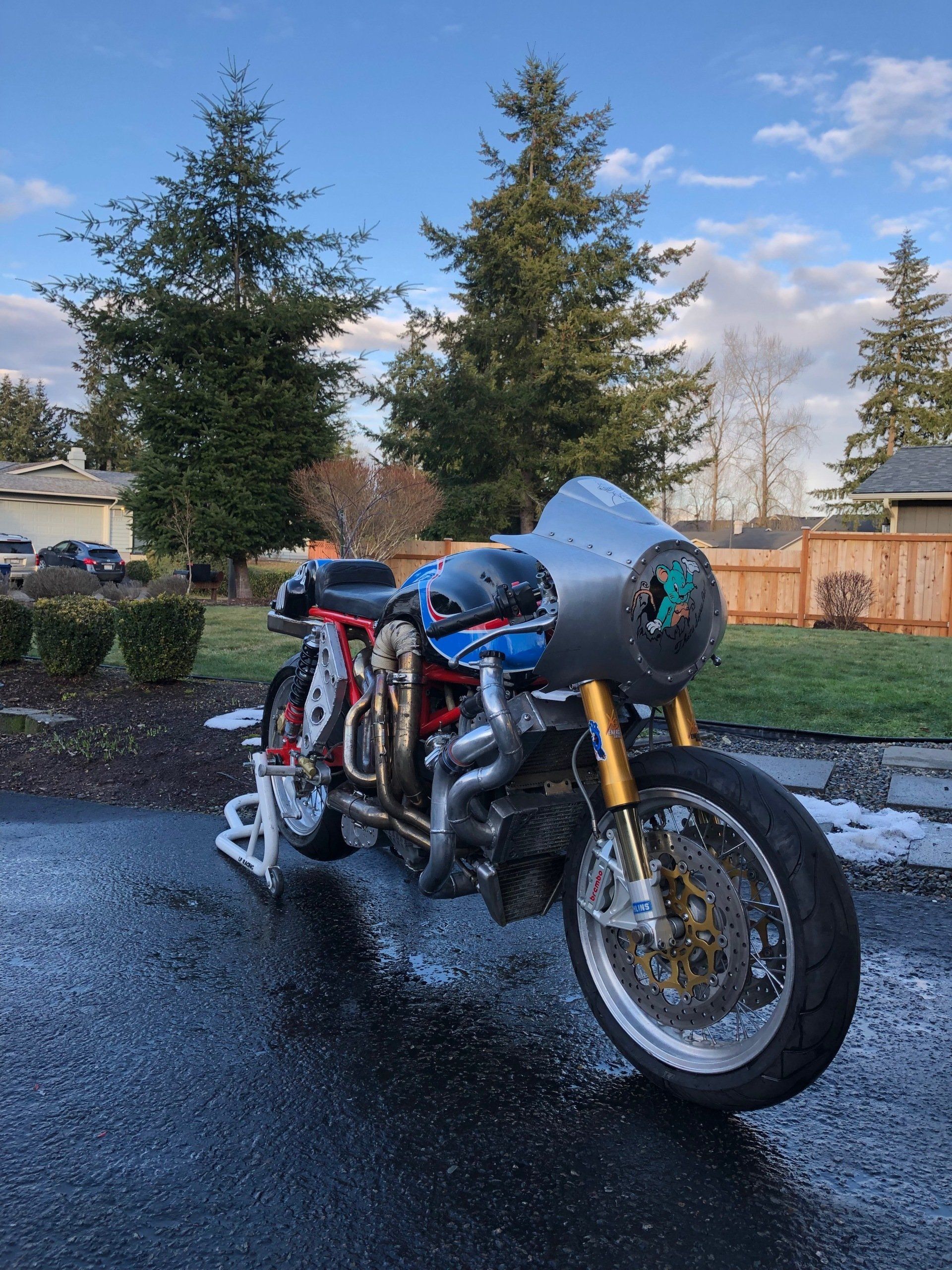 Blue motorcycle parked on wet pavement in a driveway, with trees and houses in the background