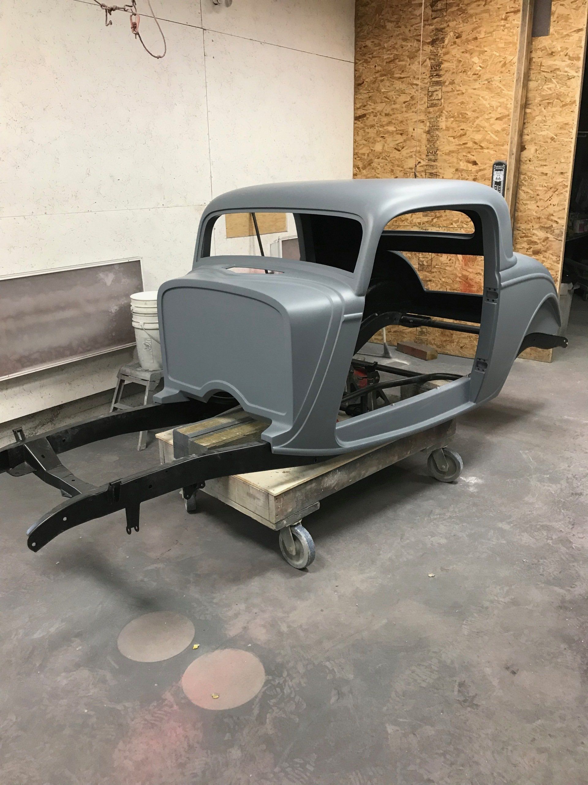 Gray vintage car body shell on a wheeled dolly in a workshop, missing wheels and interior.