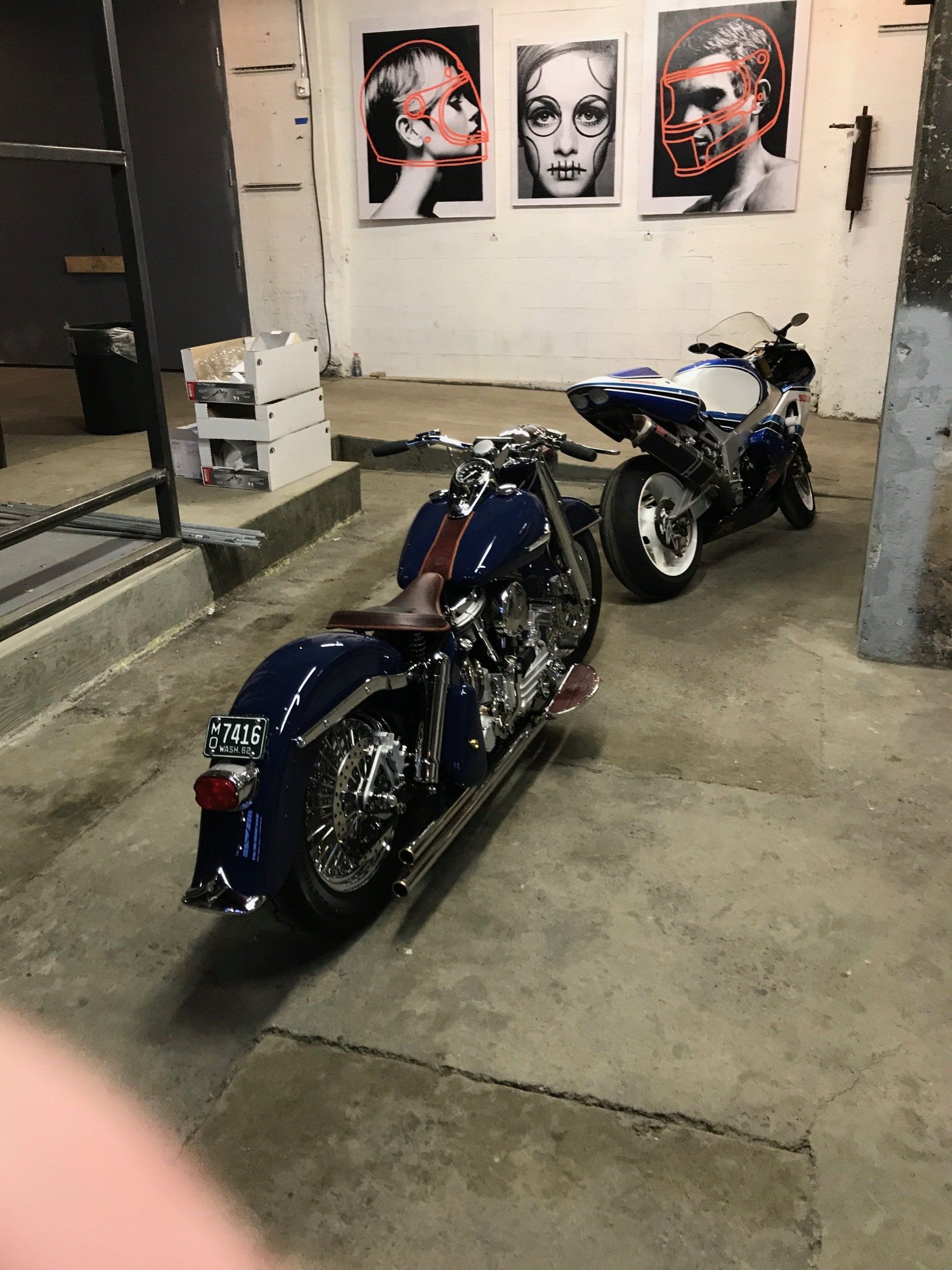 Two blue custom motorcycles displayed indoors on a concrete floor, with artwork on the wall behind them.