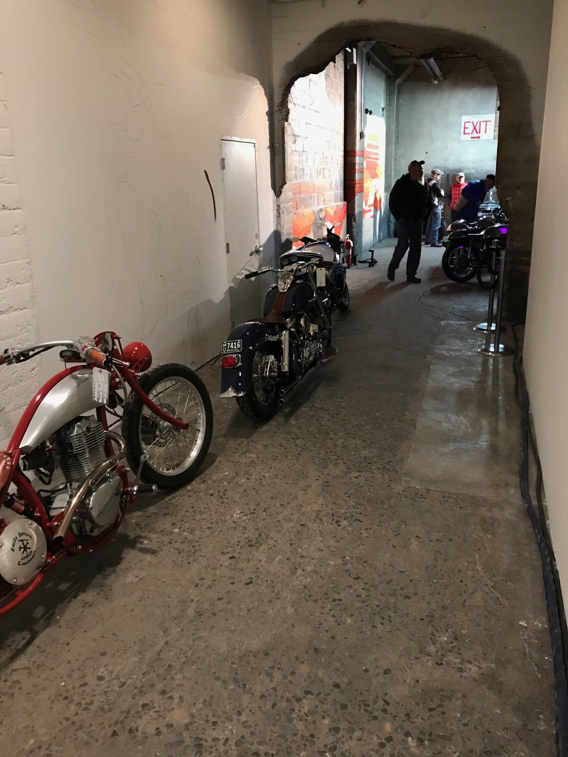 Narrow indoor hallway with motorcycles parked along the walls and two people near a lit exit sign.