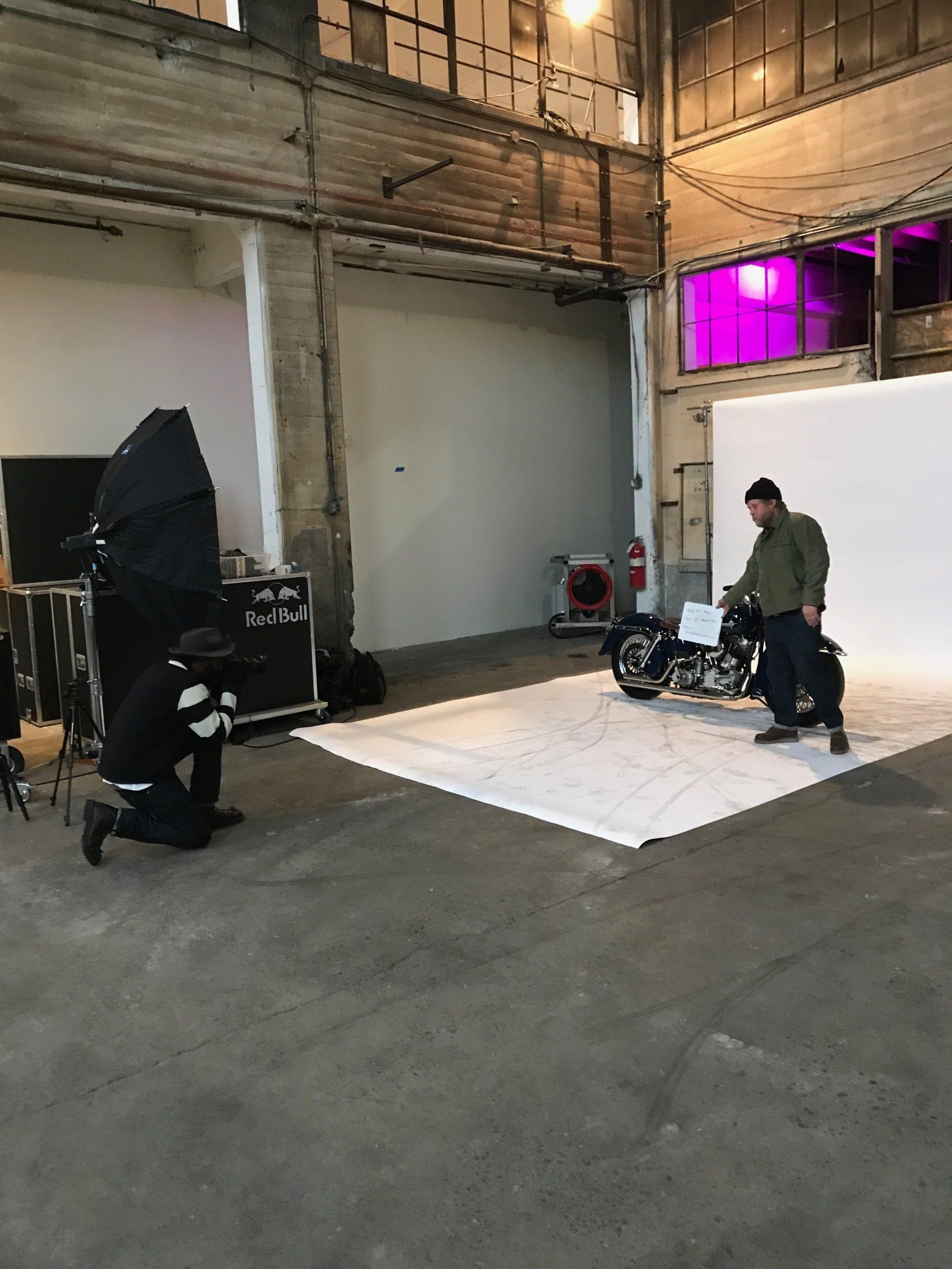 Two people adjust a white floor panel in a warehouse studio with industrial walls and purple-lit windows