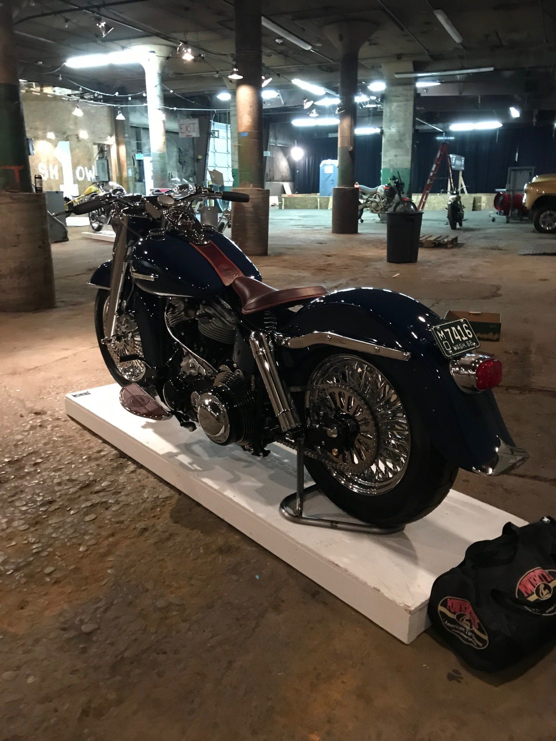 Black custom motorcycle displayed on a white platform in a dim indoor garage or showroom