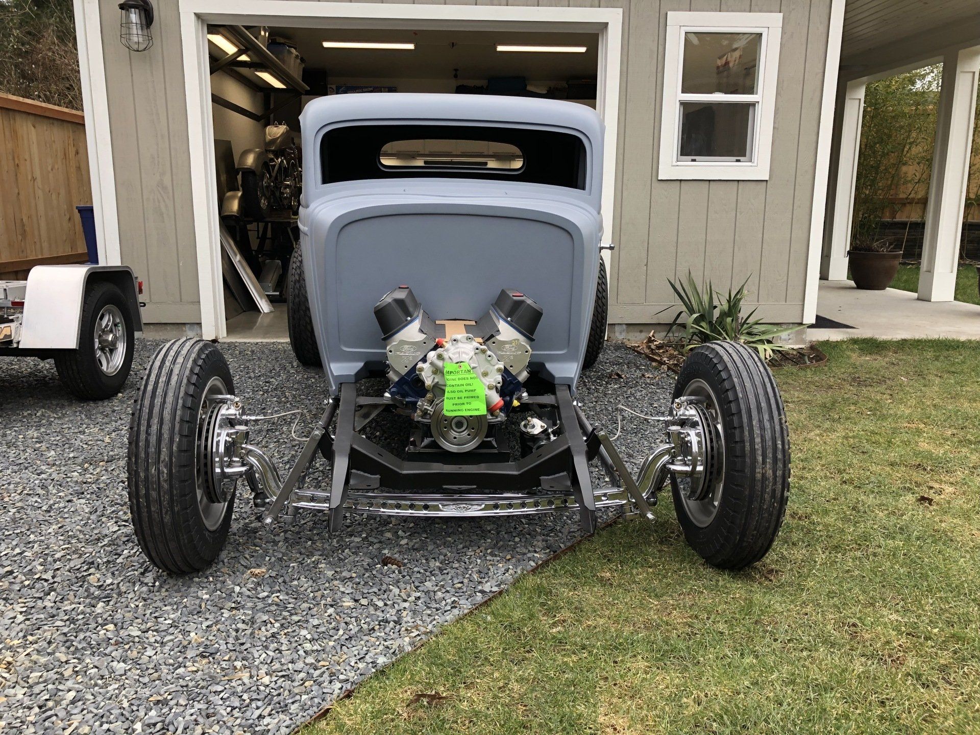 Gray vintage hot rod with exposed engine parked on gravel outside a garage.