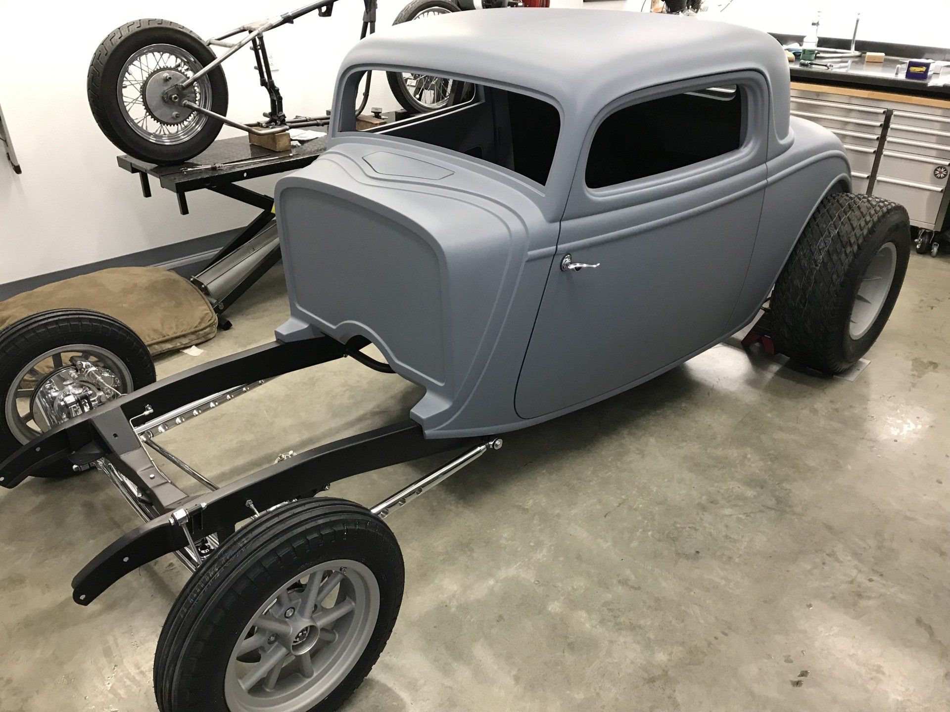 Gray vintage hot rod coupe on a car lift in a garage, with exposed front wheels and chassis.