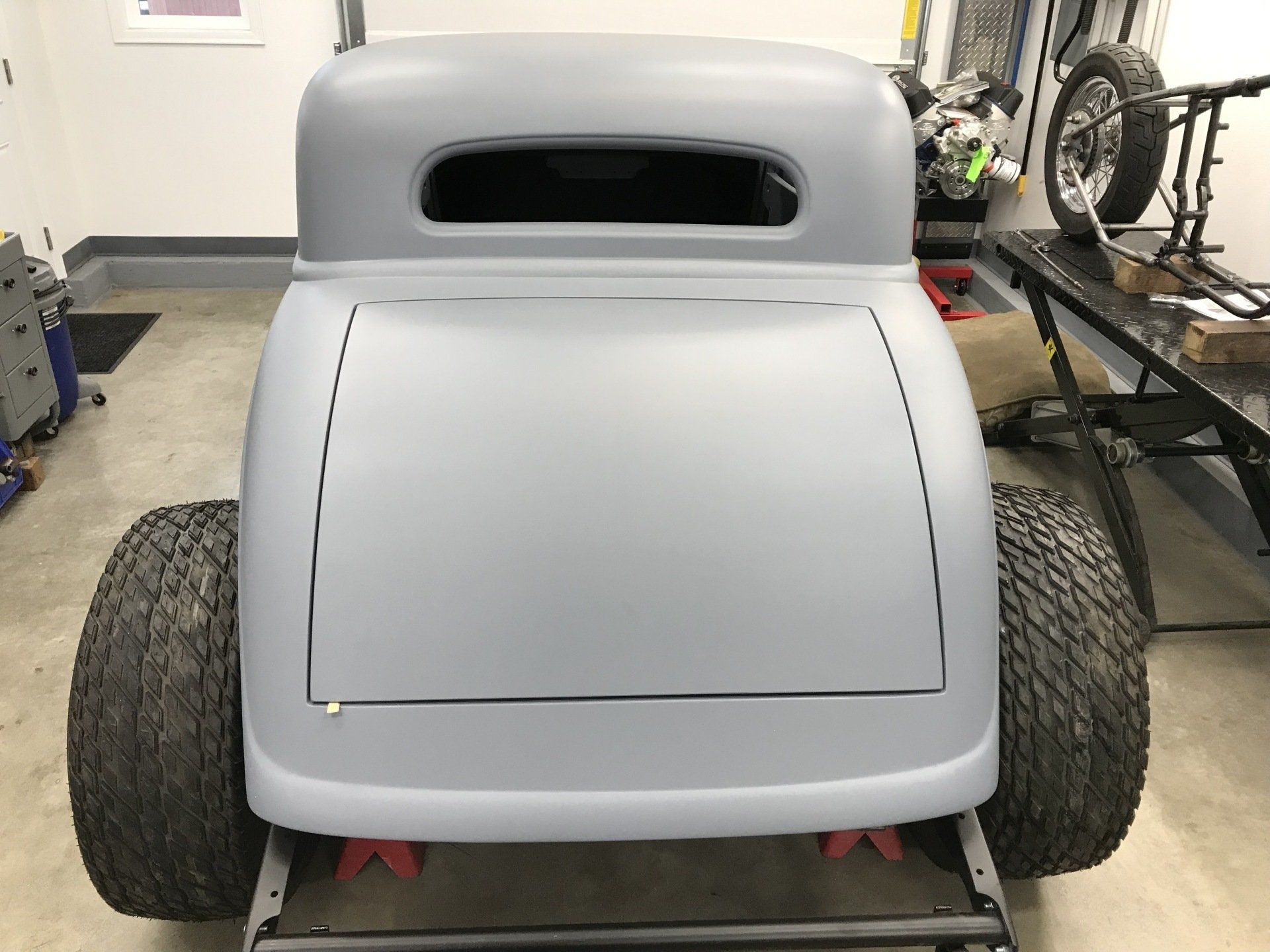 Front view of a gray custom hot rod in a garage, with wide rear tires and an open windshield slot