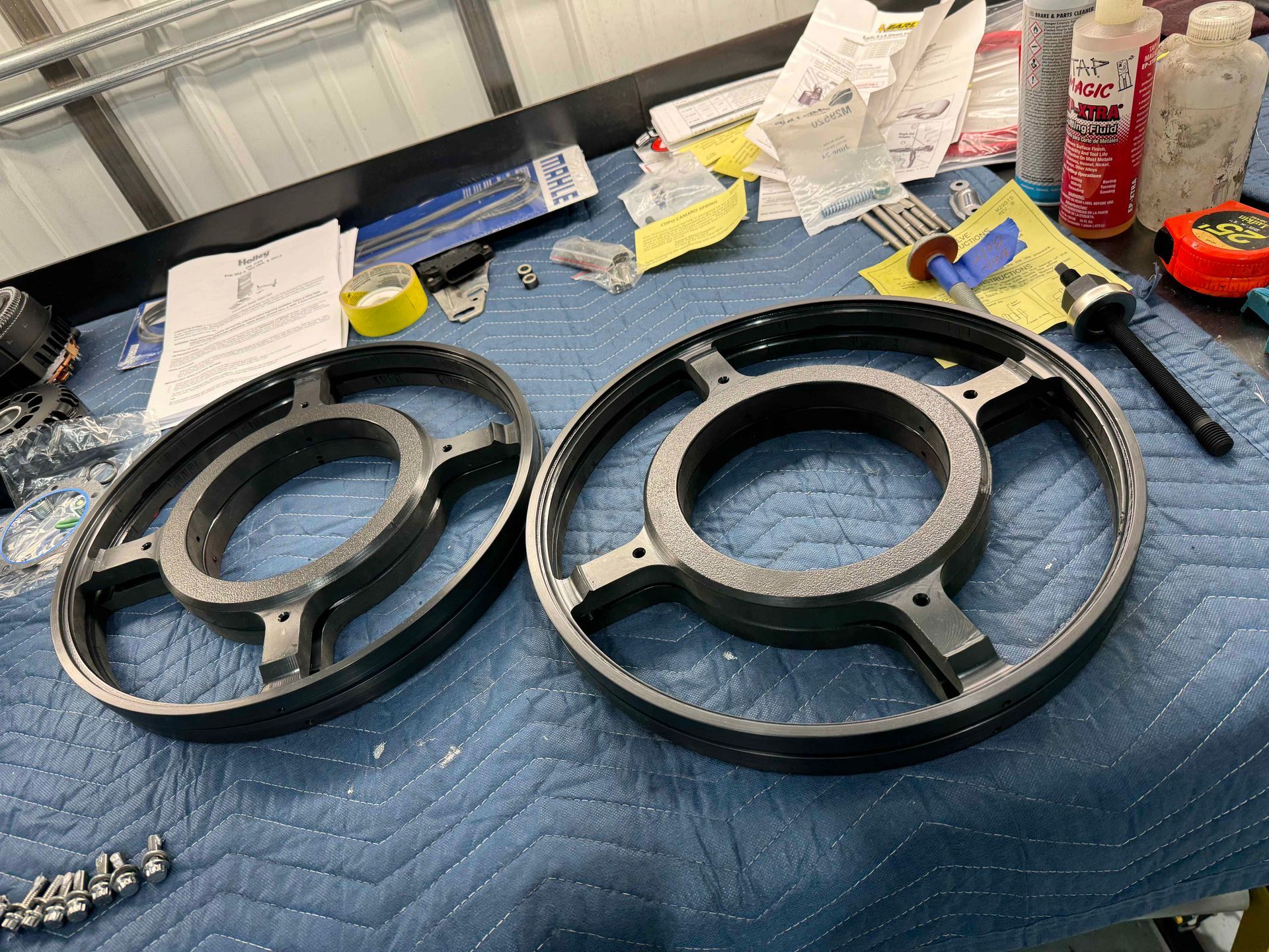 Two black circular metal parts on a blue quilted mat, with tools and supplies scattered on a workbench.