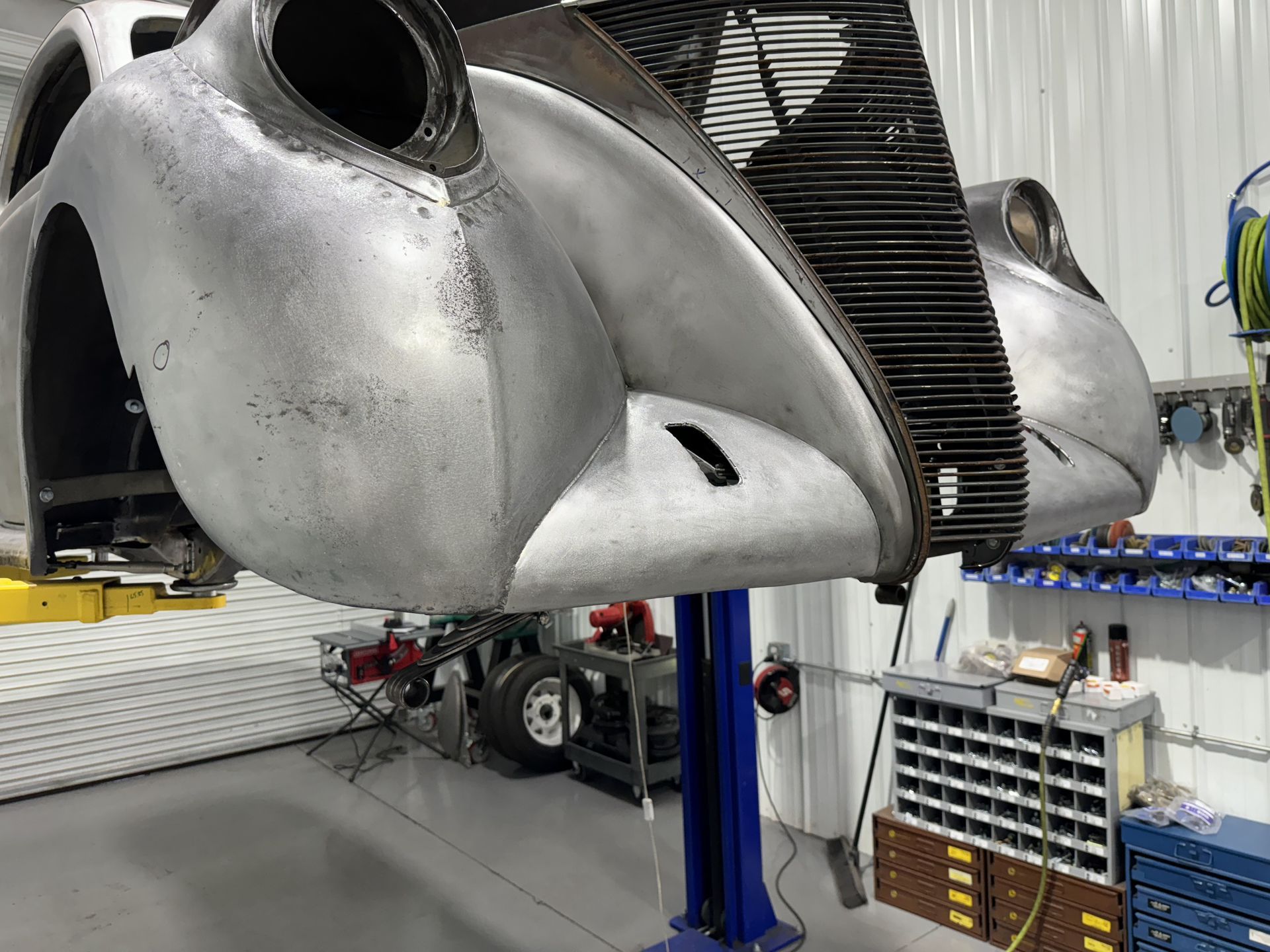 Gray aircraft fuselage under repair in a workshop, mounted on a blue lift with tools and shelves nearby