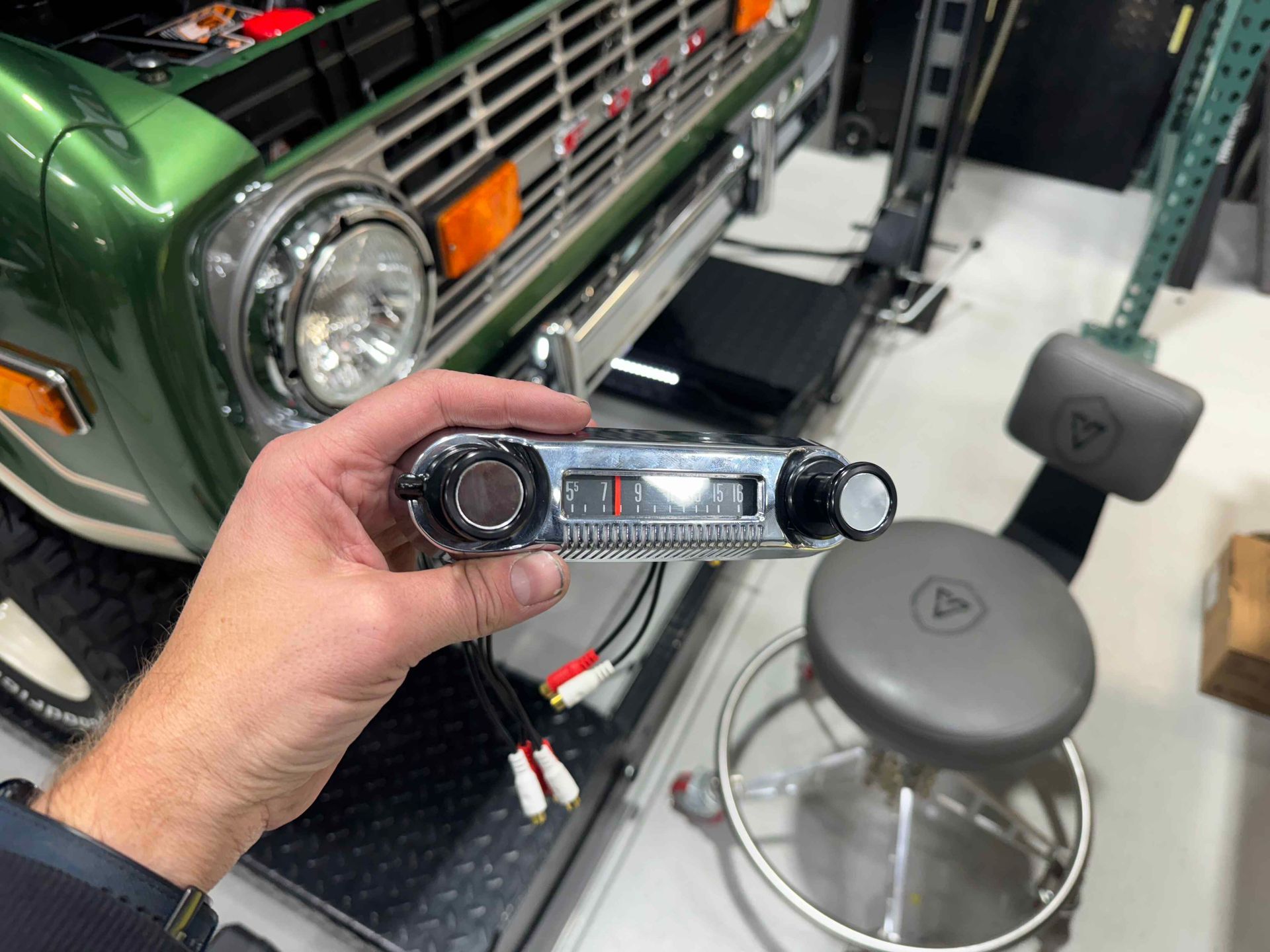 Hand holding a car stereo head unit beside a green vehicle and workshop stool in a garage