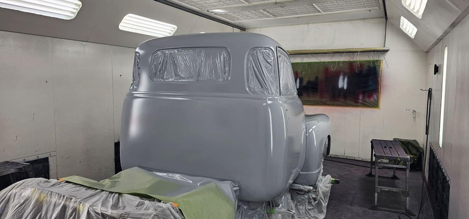 Gray armored vehicle body in a paint booth, covered in protective plastic and overspray.