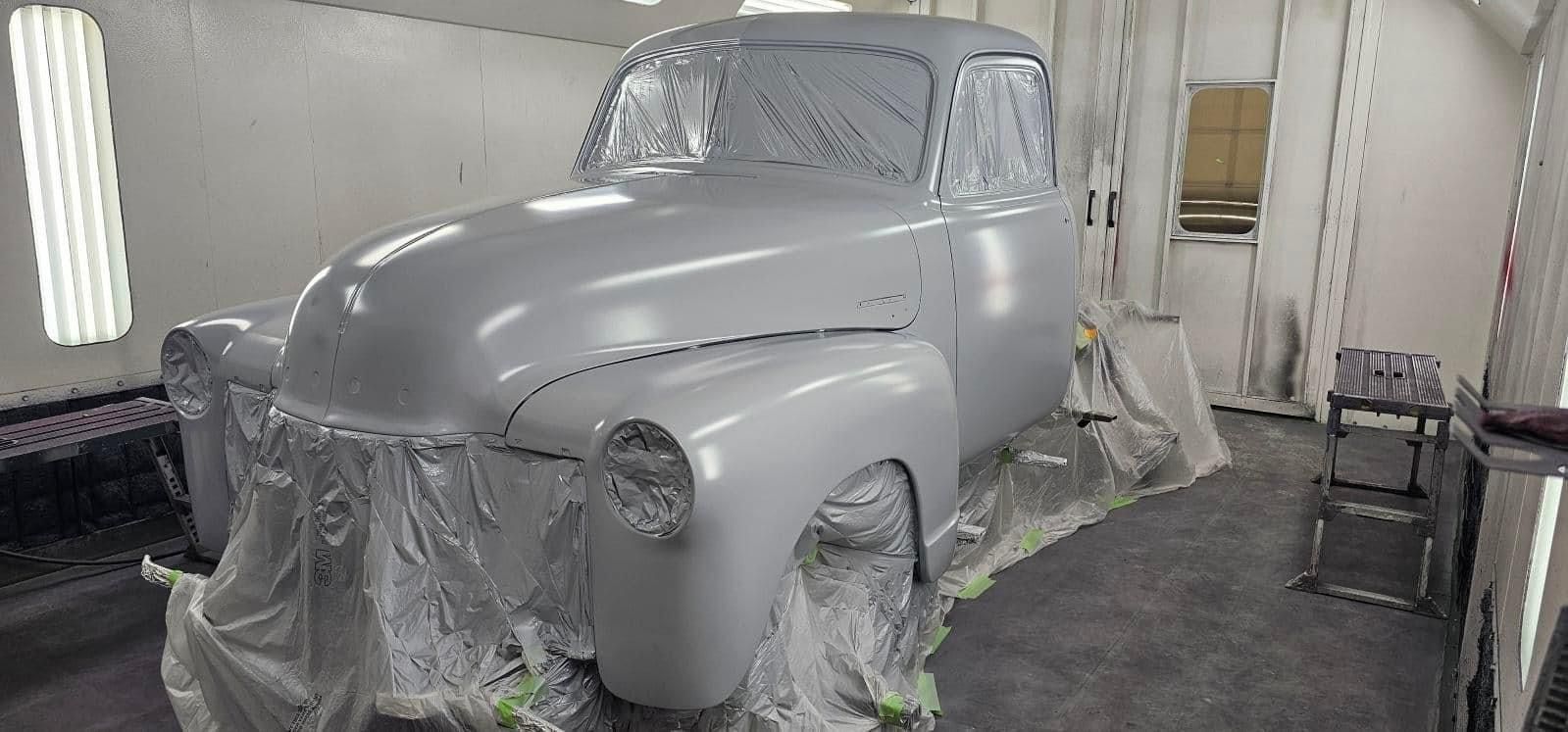 Gray vintage truck covered in masking, parked in a paint booth during restoration