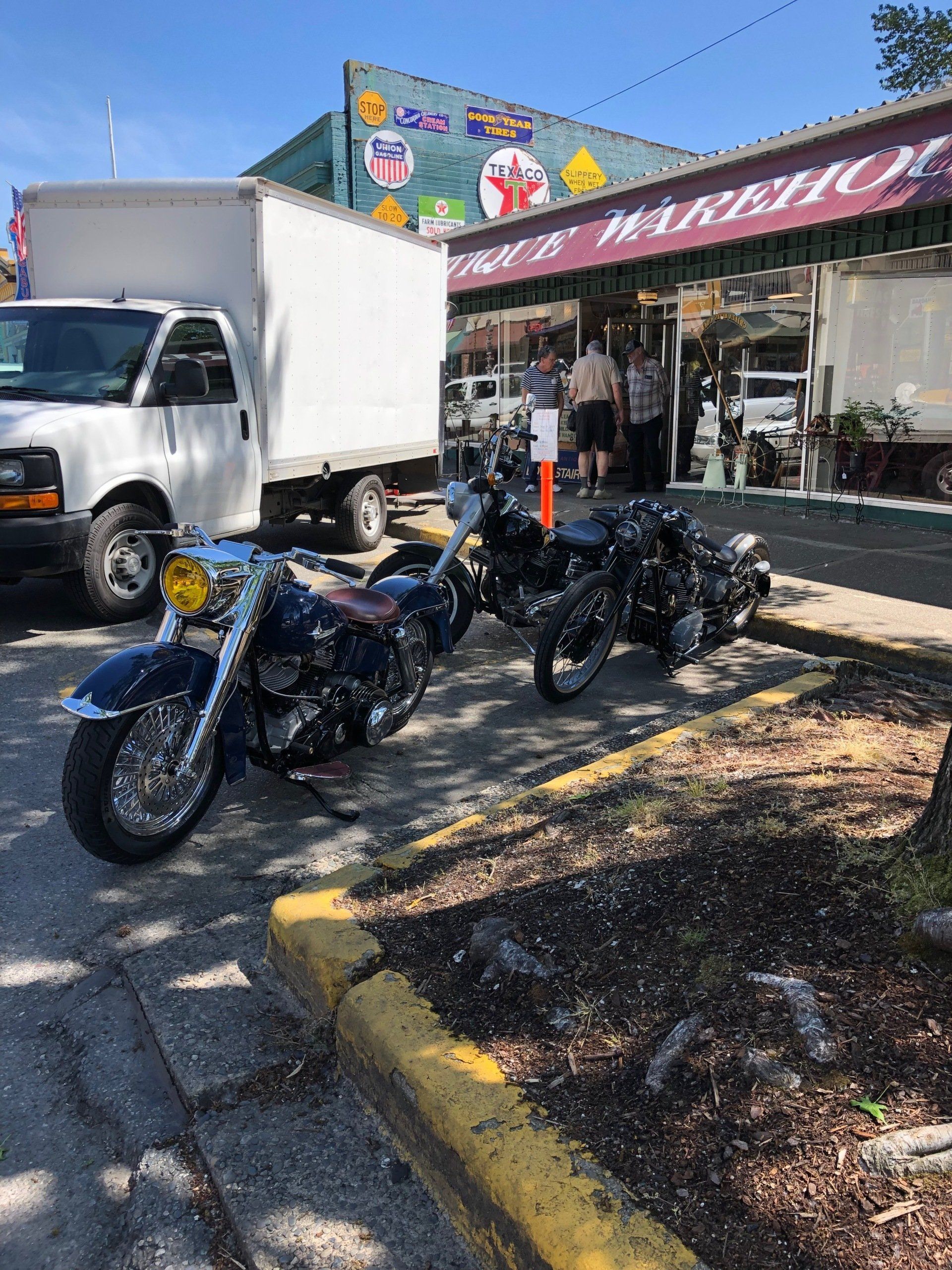 Motorcycles parked outside a store with a white truck nearby on a sunny street.