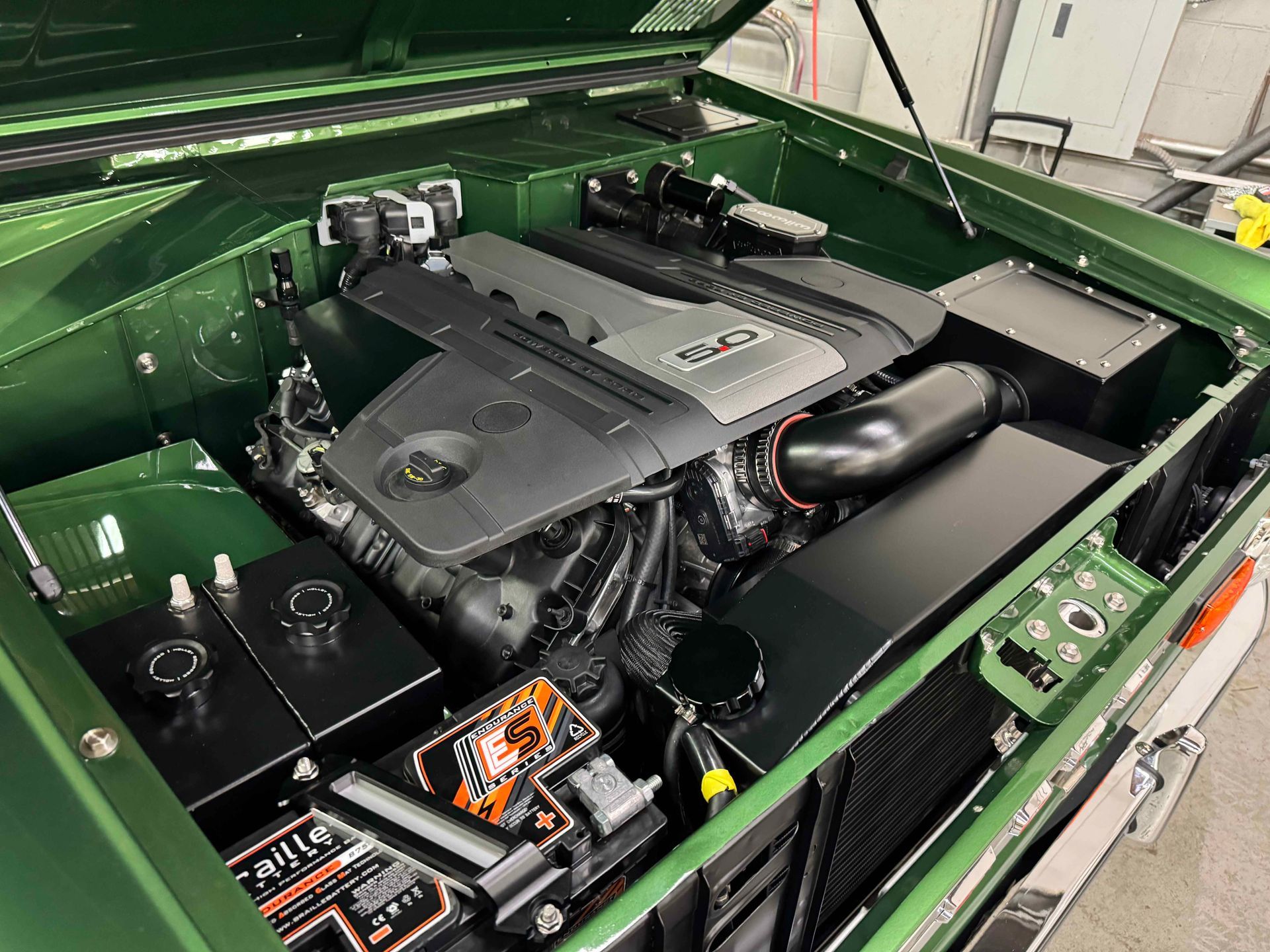 Open car hood showing a large black engine bay with green body panels and visible battery components