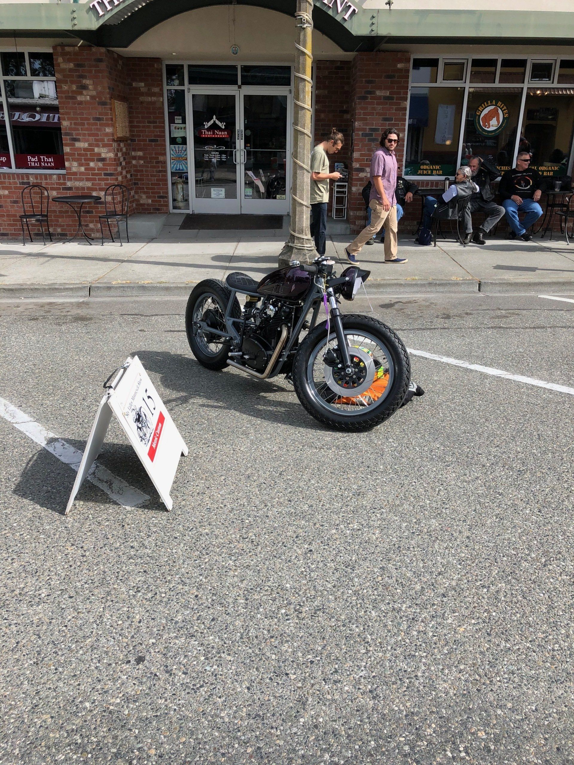 Black motorcycle on display outside a brick storefront, with a small sign in the street.