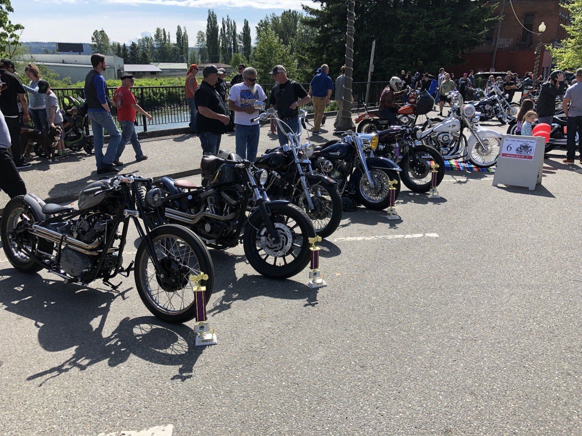 Row of custom motorcycles parked outdoors with people standing nearby on a sunny day