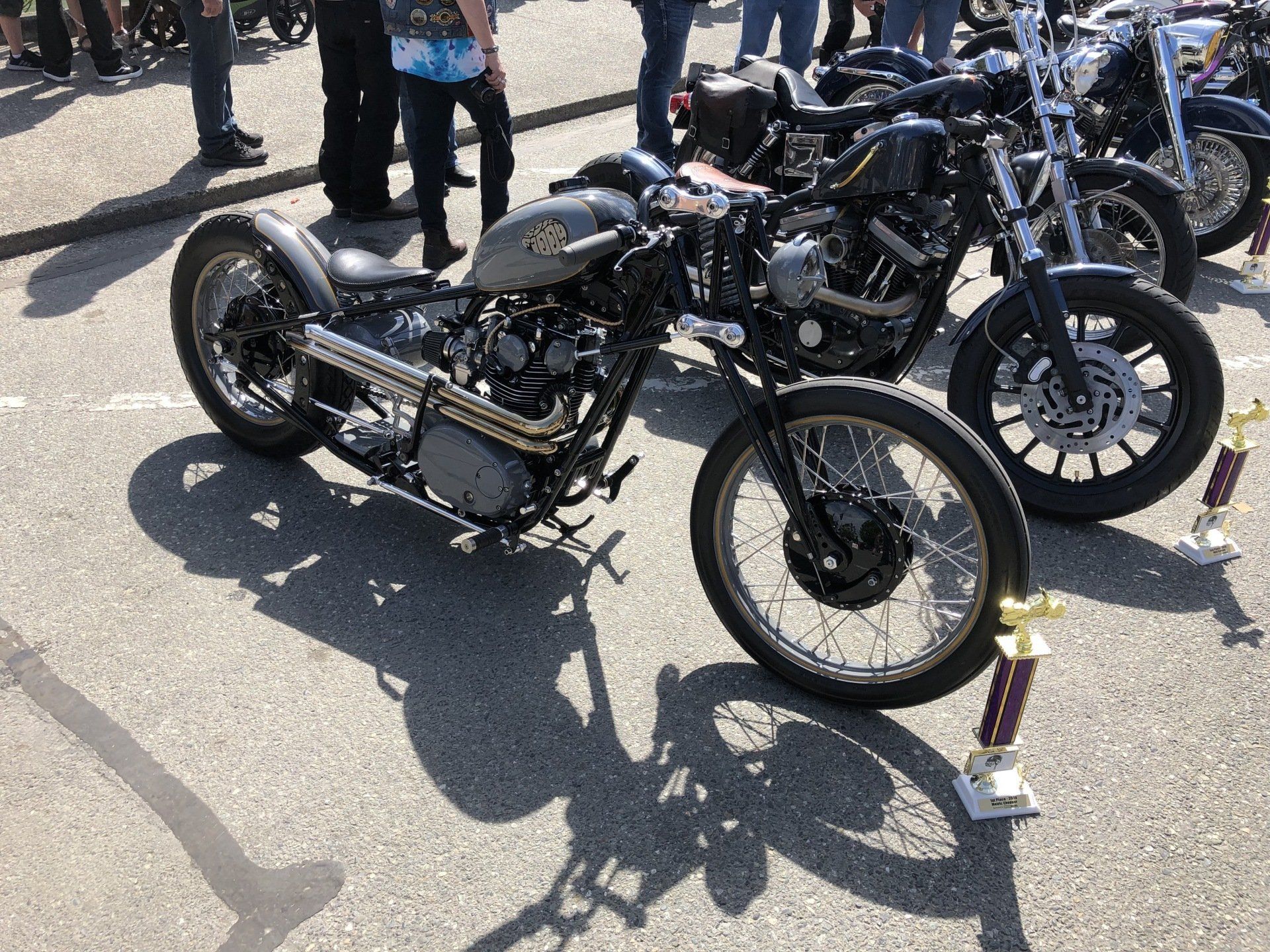 Two custom black motorcycles parked on pavement, with people standing in the background.