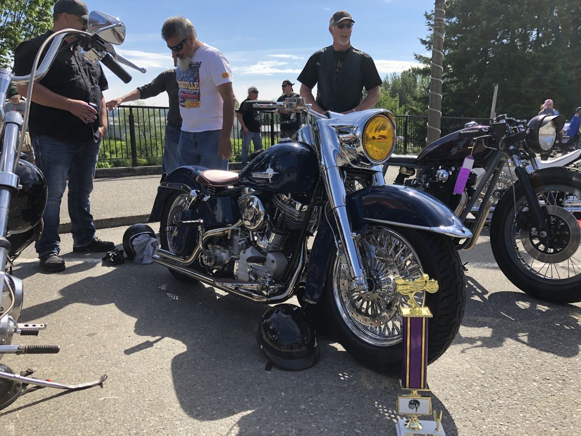 Motorcycles and riders gathered outdoors, with a blue bike and trophies in the foreground.