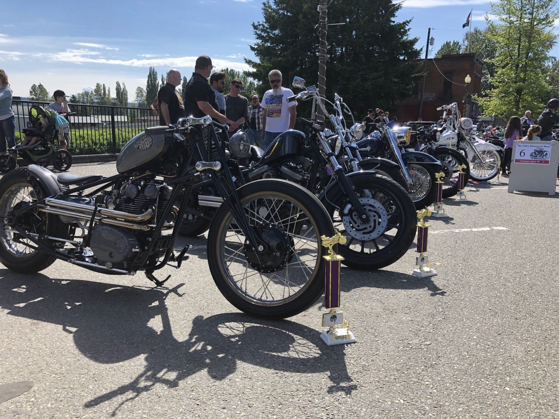 Row of black custom motorcycles at an outdoor show with people standing nearby in bright sunlight