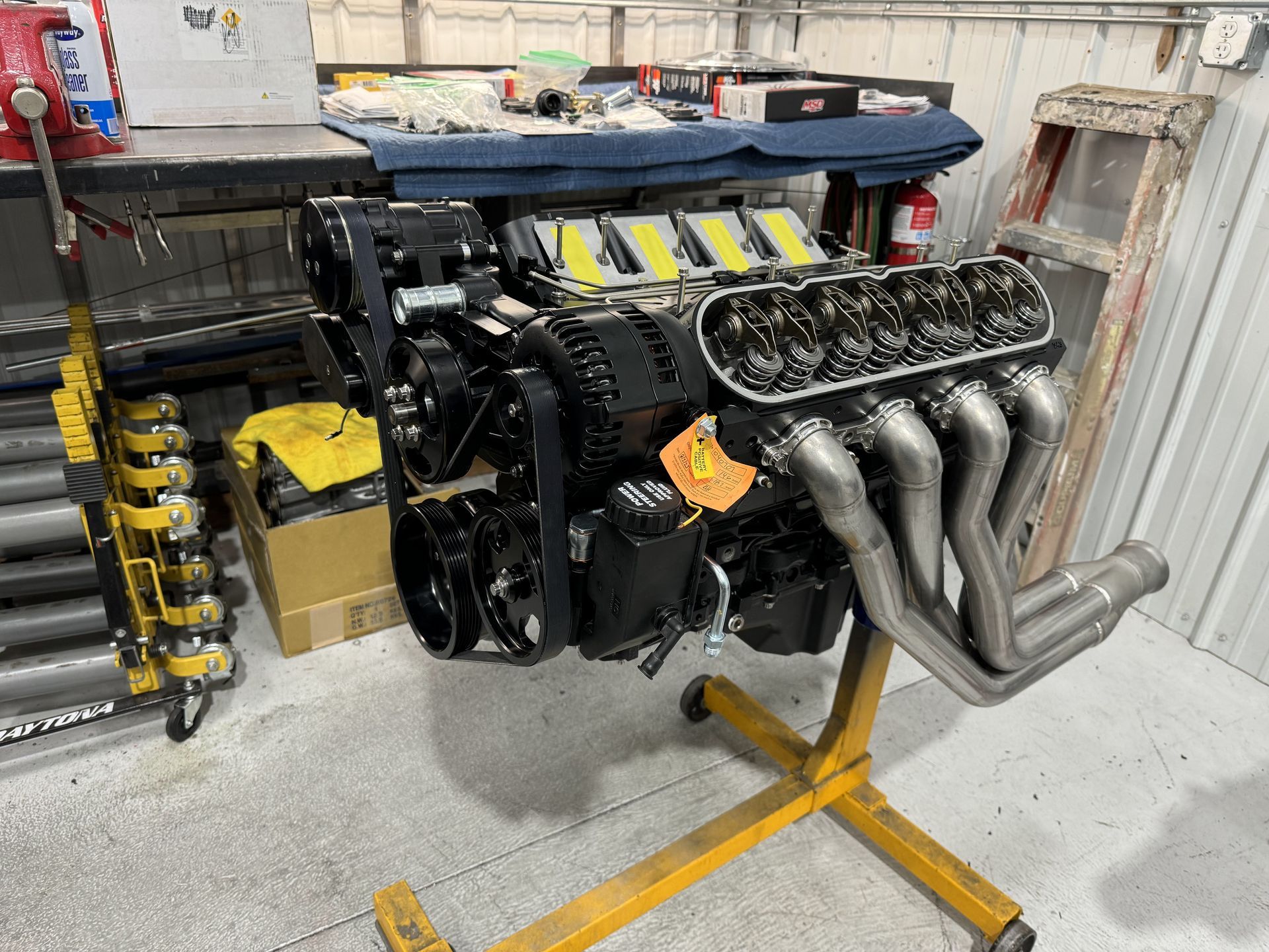 Engine on a yellow stand in a workshop, with exposed exhaust headers and yellow/black components.