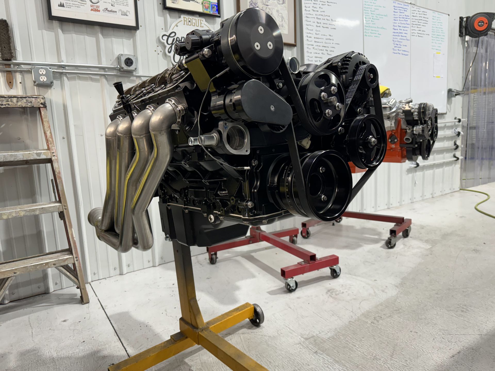 Large black engine on a red stand in a workshop, with exhaust headers and exposed pulleys.