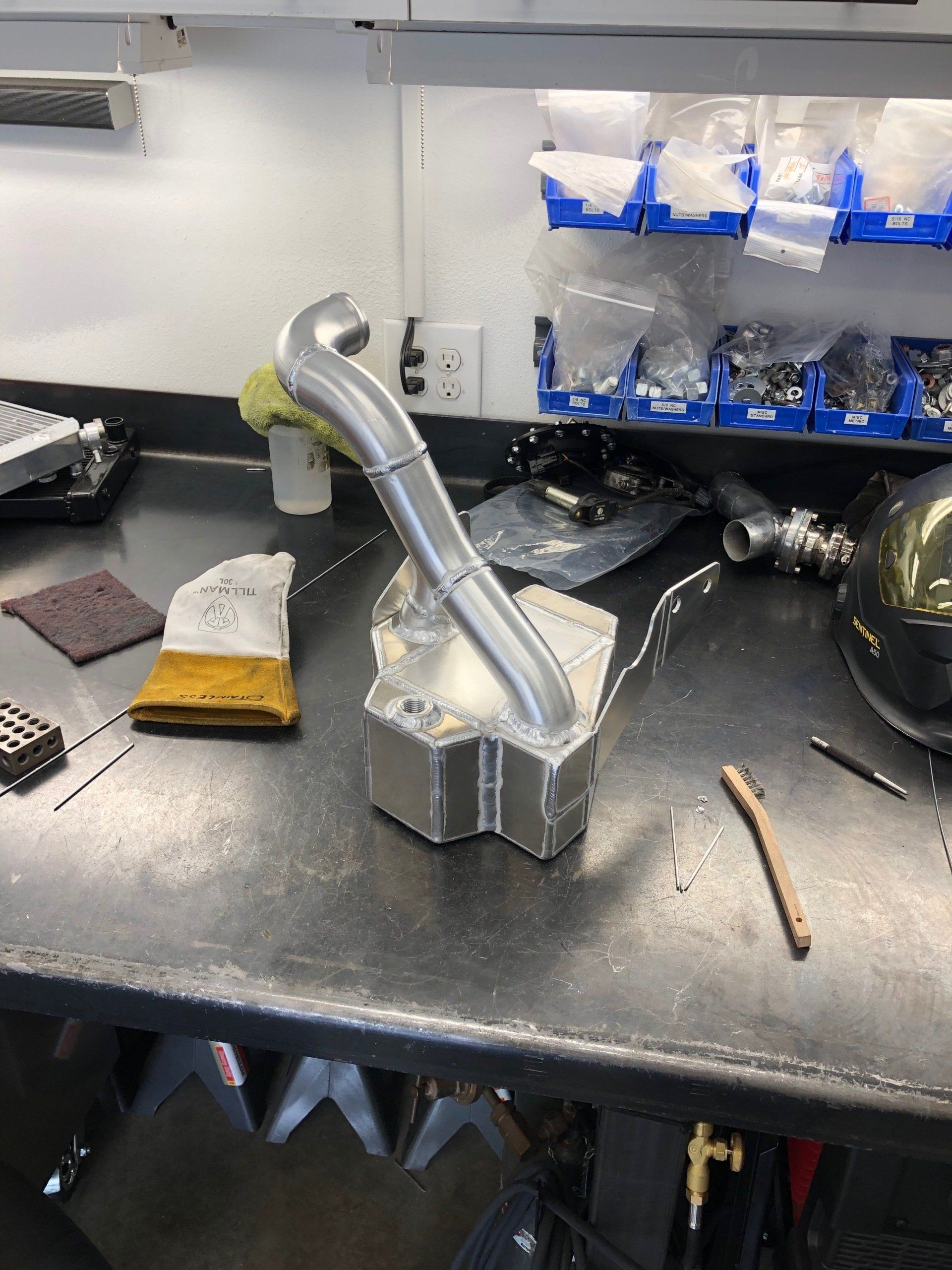 Workshop bench with a metal pipe fitting prototype in a vise, surrounded by tools and parts
