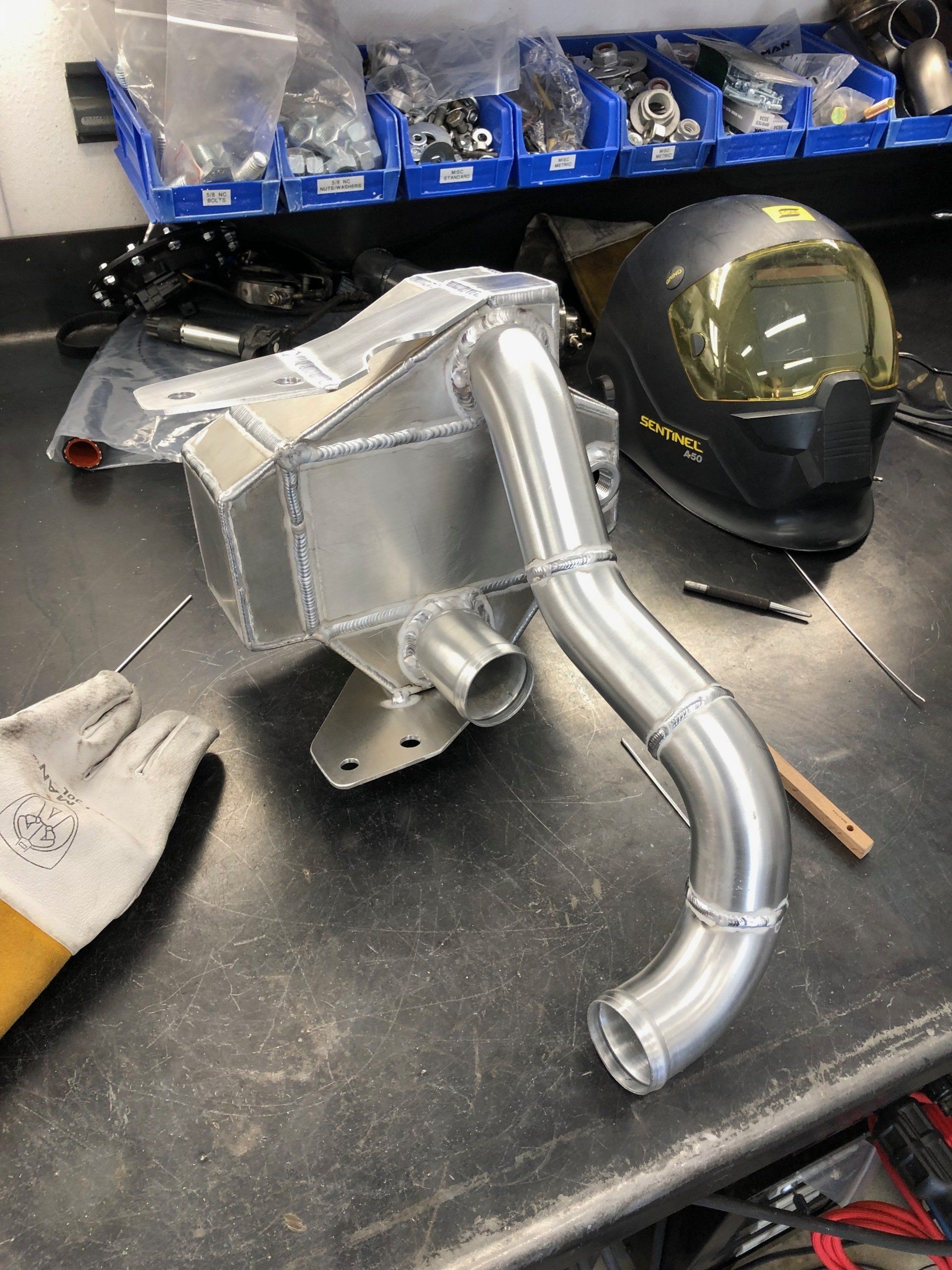 Metal automotive part on a workbench beside a welding helmet and tools in a workshop.
