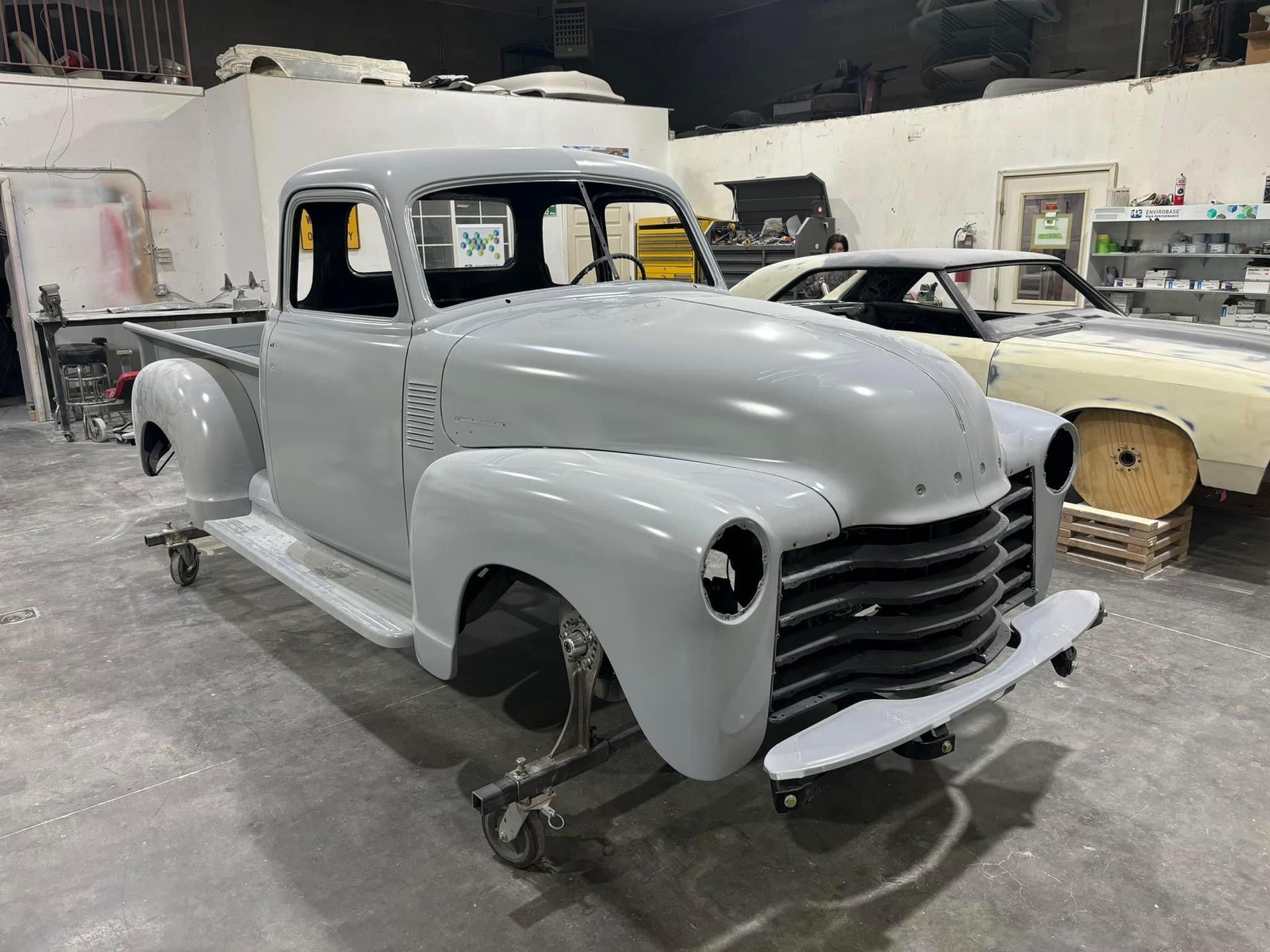 Gray vintage pickup truck body in a workshop, missing wheels and parts, with other classic cars nearby