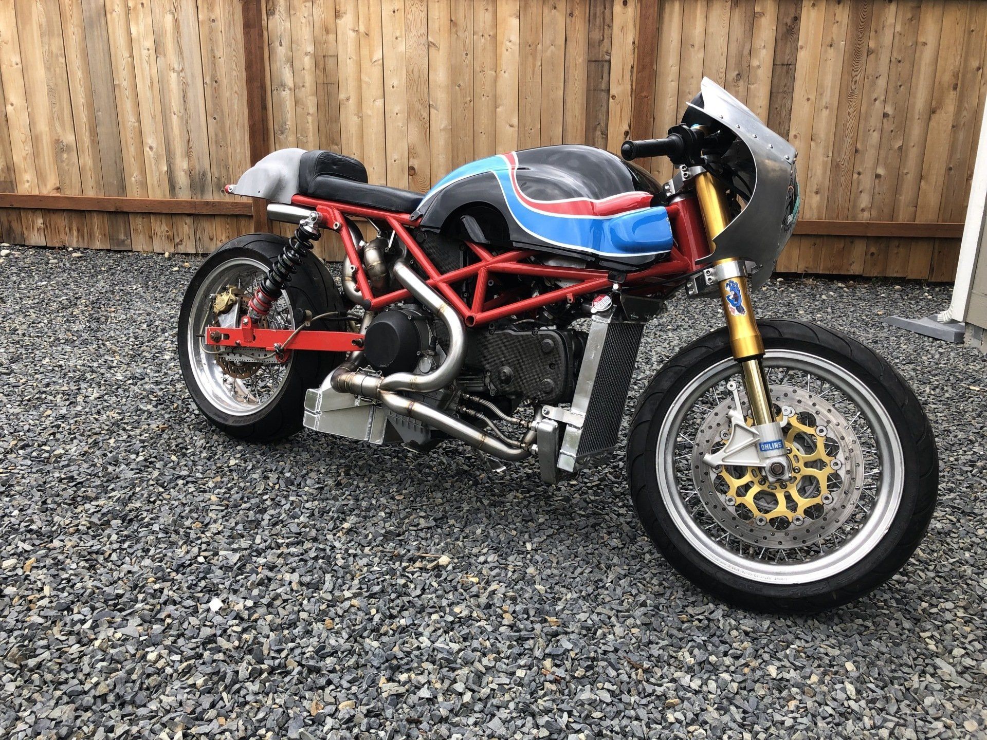 Red custom sport motorcycle with blue fuel tank parked on gravel in front of a wooden fence.