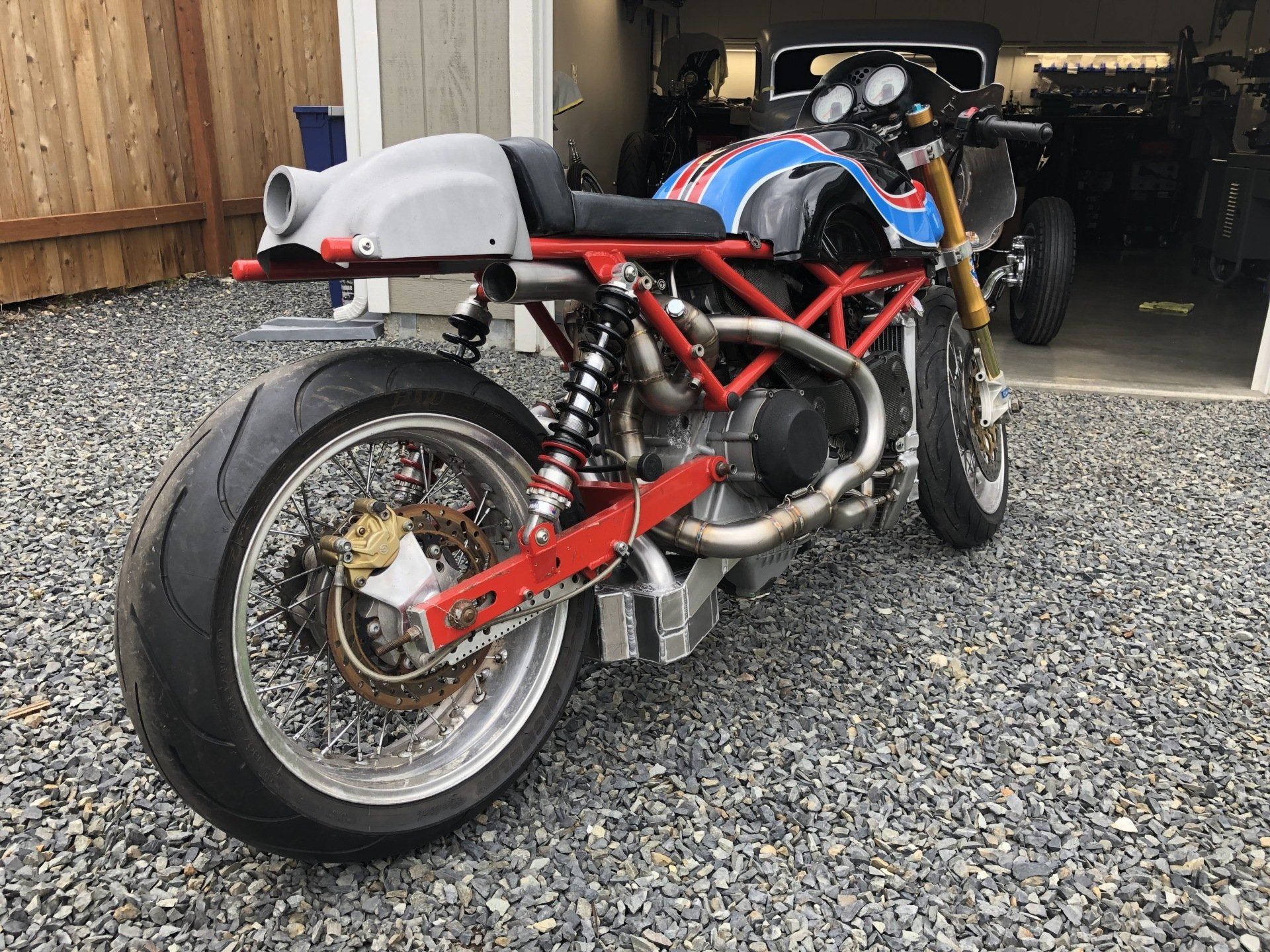 Custom red sport motorcycle parked on gravel in front of a garage, rear three-quarter view