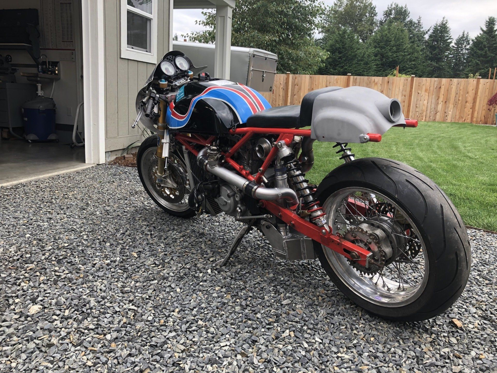 Custom blue-and-red sport motorcycle parked on gravel beside a house and wooden fence