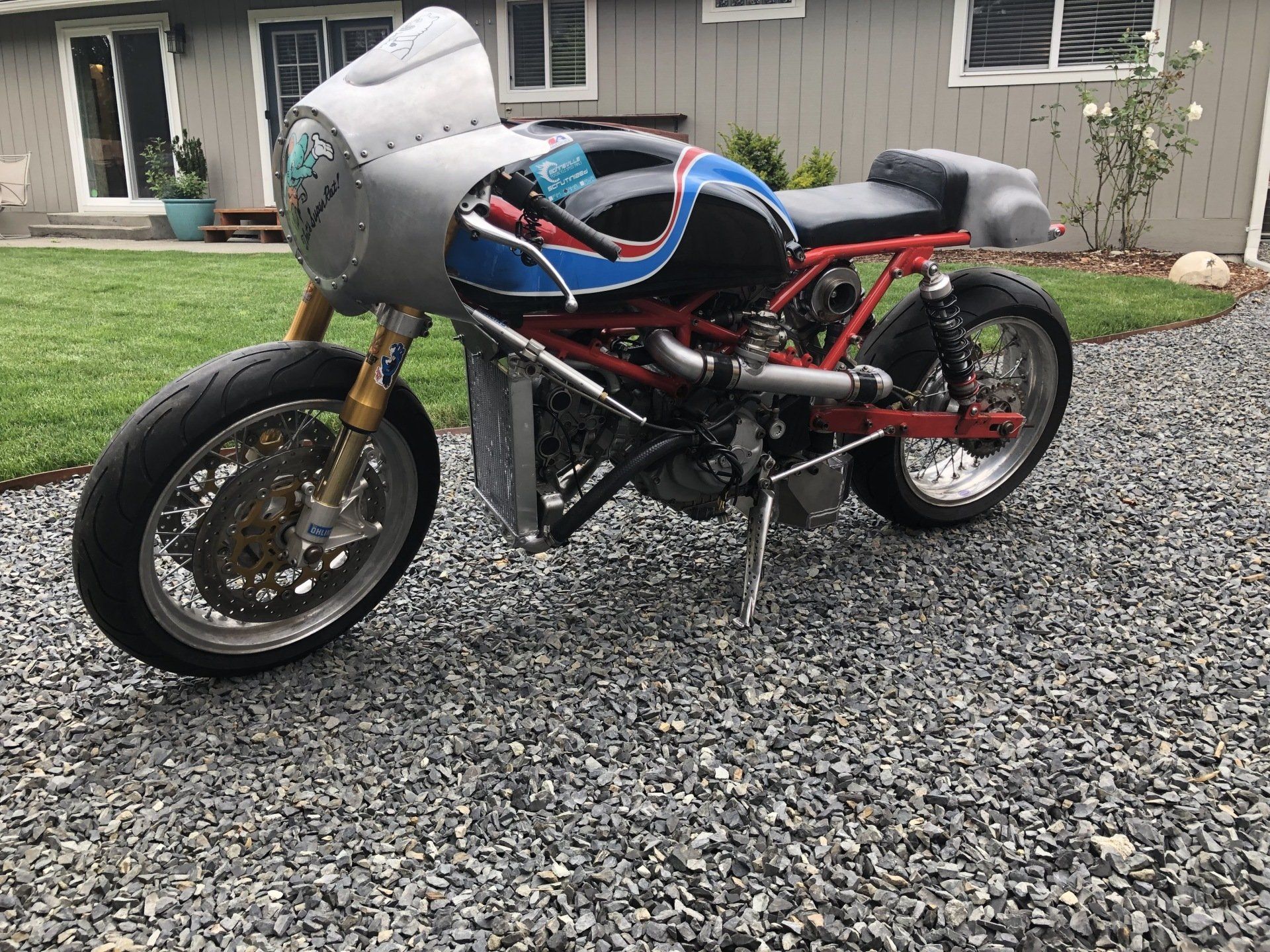 Custom racing motorcycle with silver fairing and red frame parked on gravel in a yard