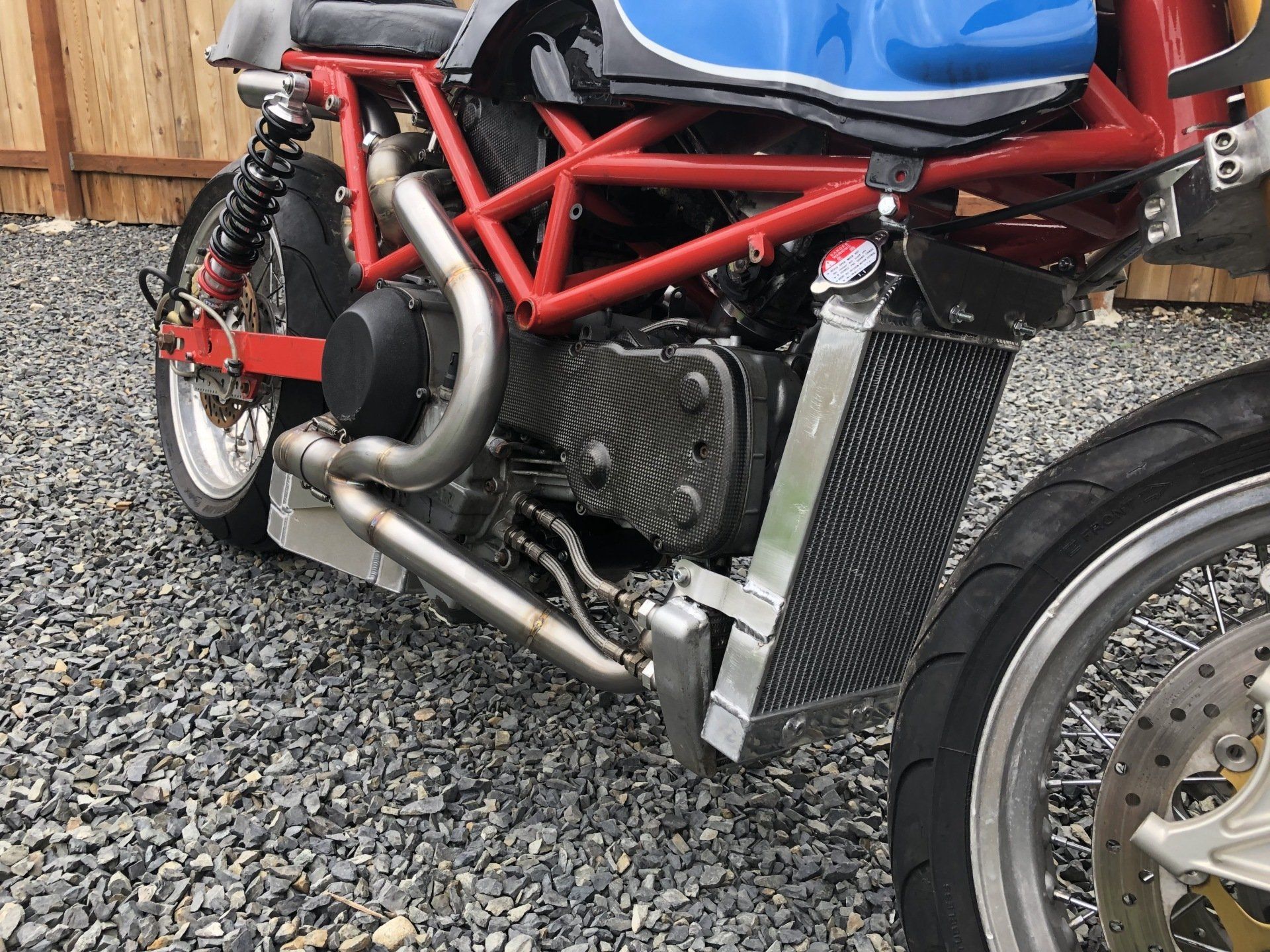Blue and red custom motorcycle with exposed engine, parked on gravel beside a wooden fence.