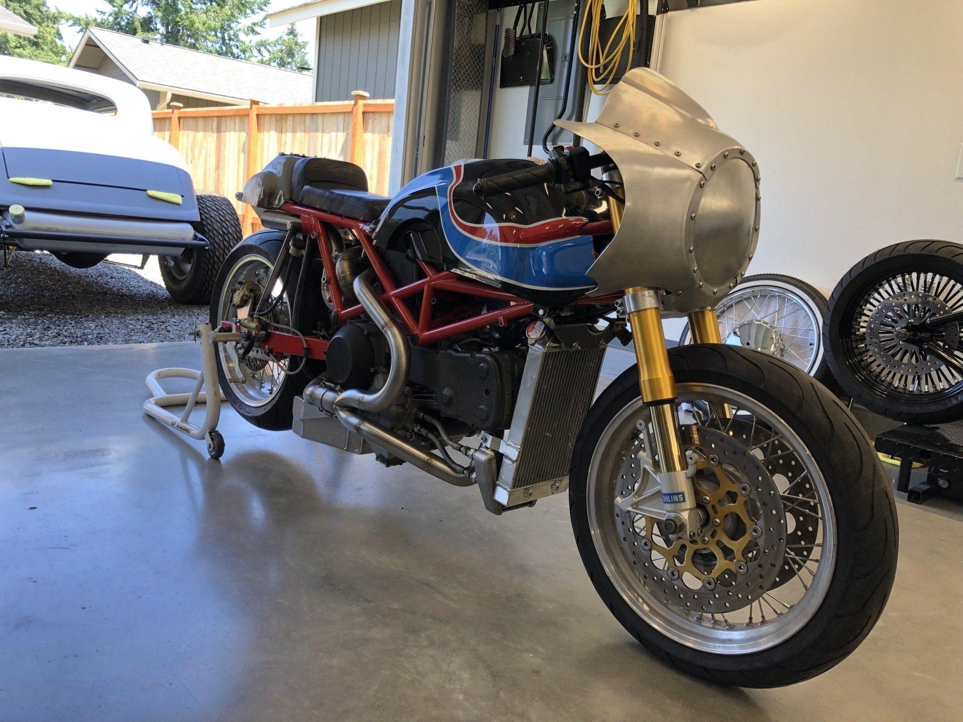 Custom motorcycle with red frame and blue tank in a garage, parked beside a white car.