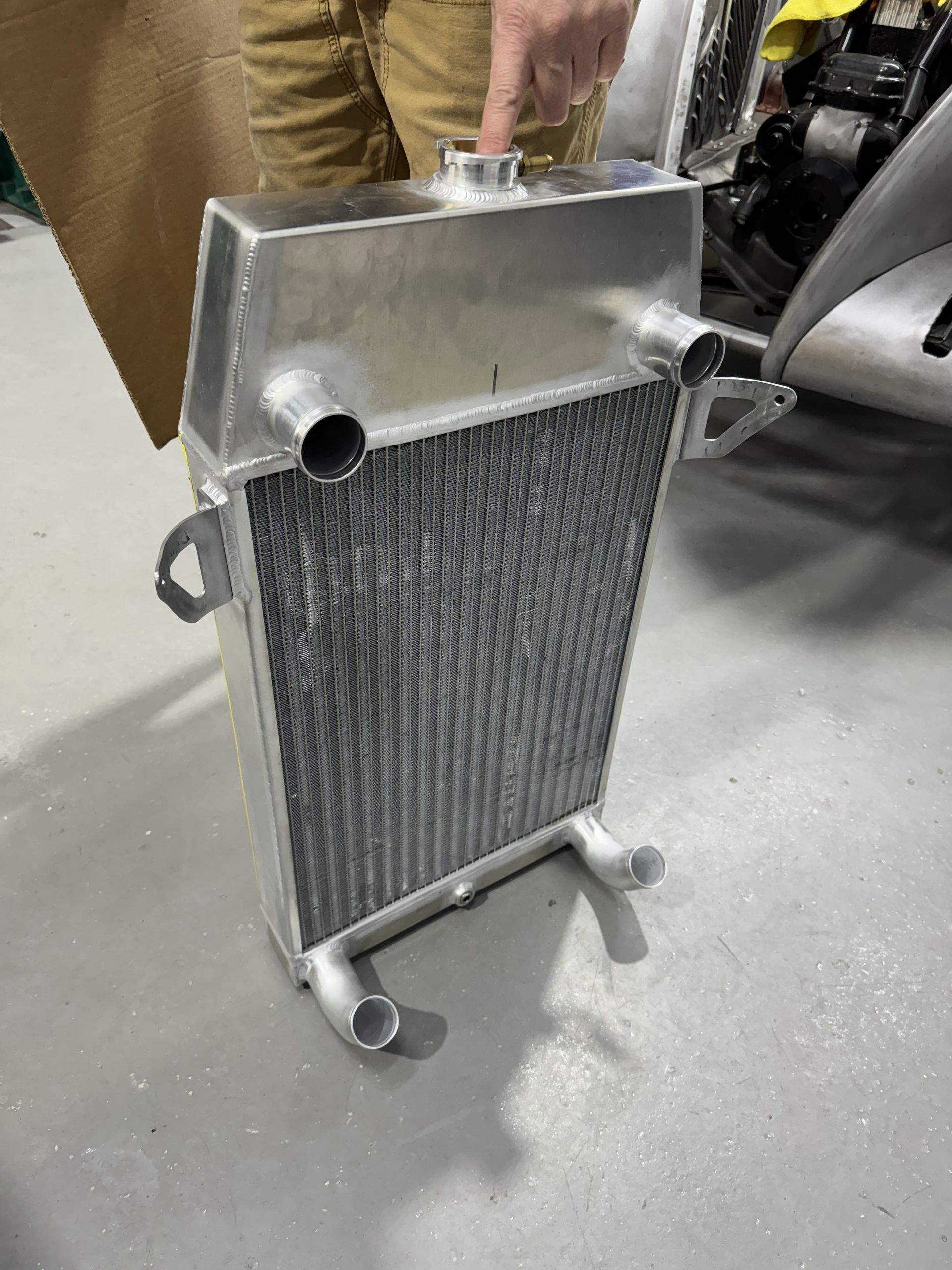 Metal car radiator with attached pipes standing upright on a workshop floor, held by a person’s hand.