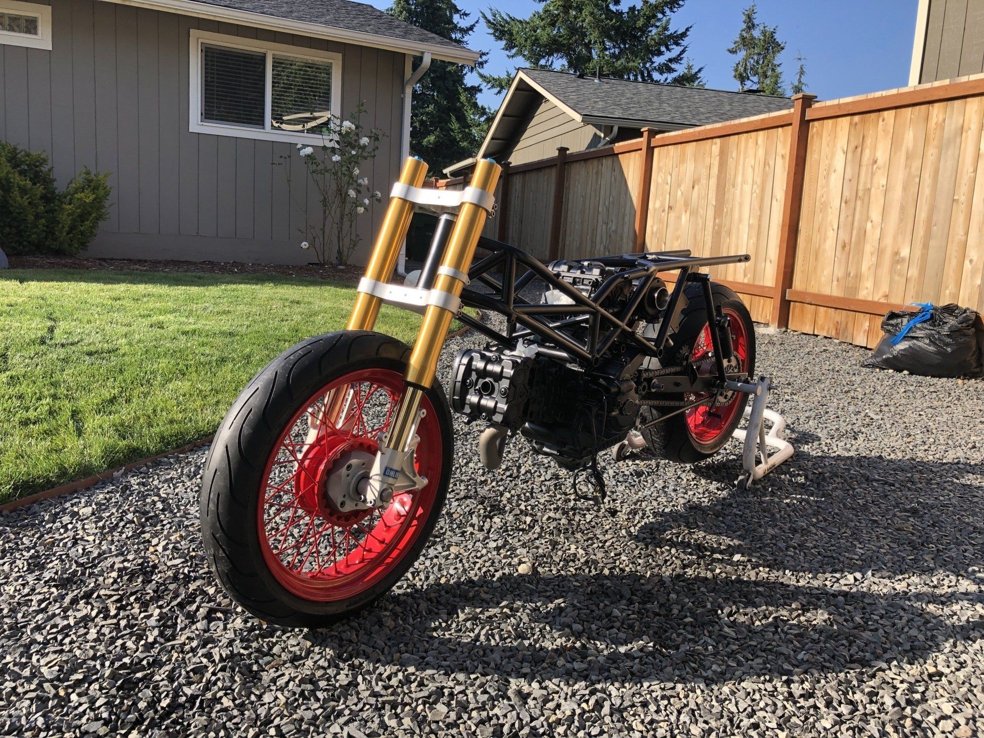 Custom black motorcycle with red wheels parked on gravel beside a wooden fence and house.