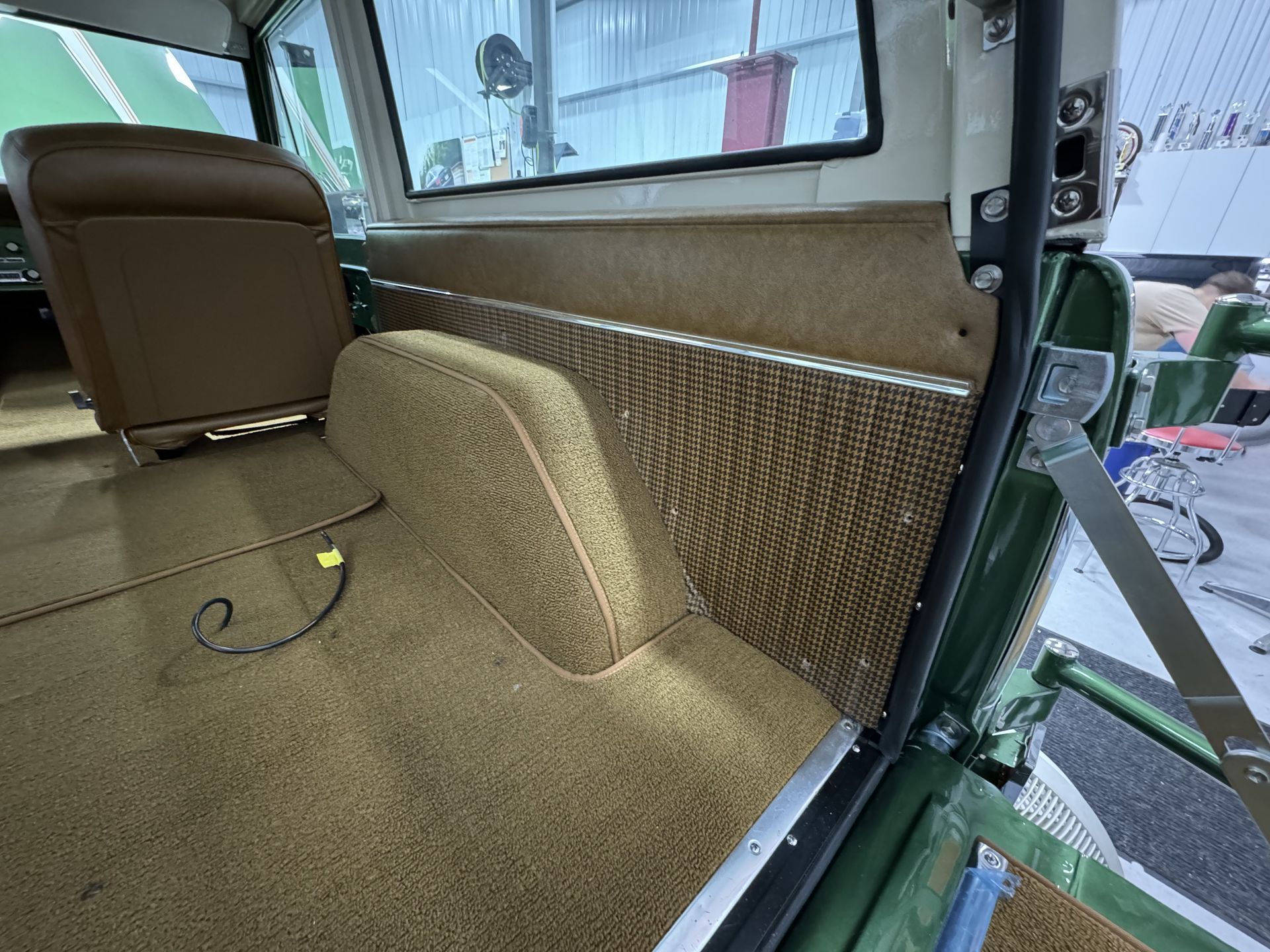Carpeted vehicle interior with tan bench seat, woven side panel, and open door showing outside.