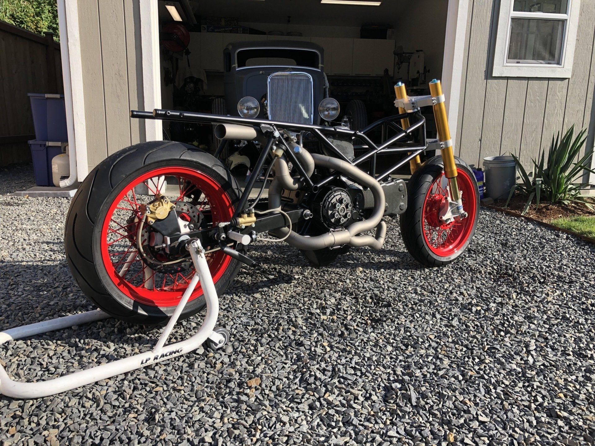 Custom motorcycle with red rims parked on gravel outside a garage, with a hose attached to the front wheel.
