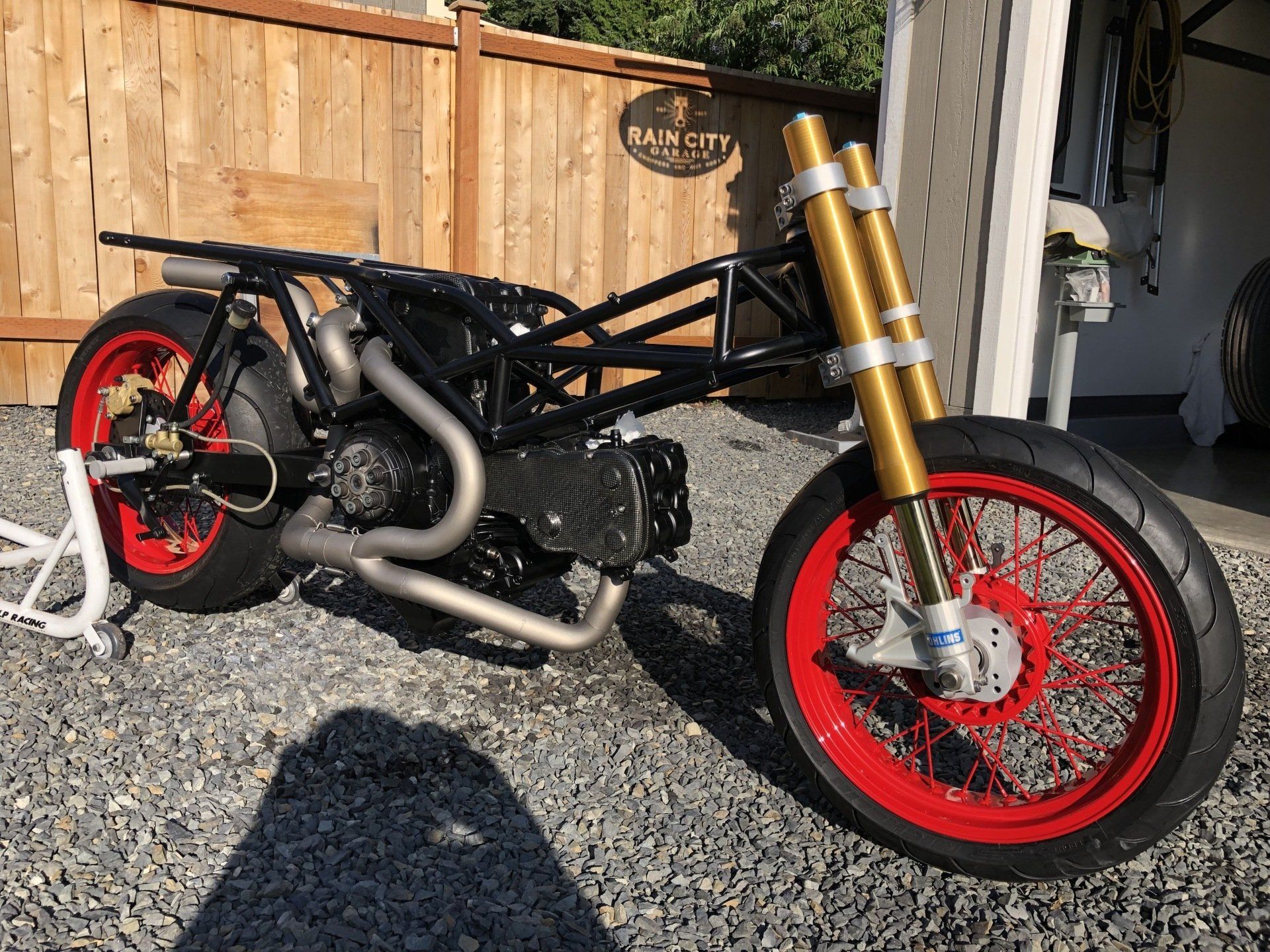 Custom black motorcycle with red wheels parked on gravel beside a wooden fence and garage.