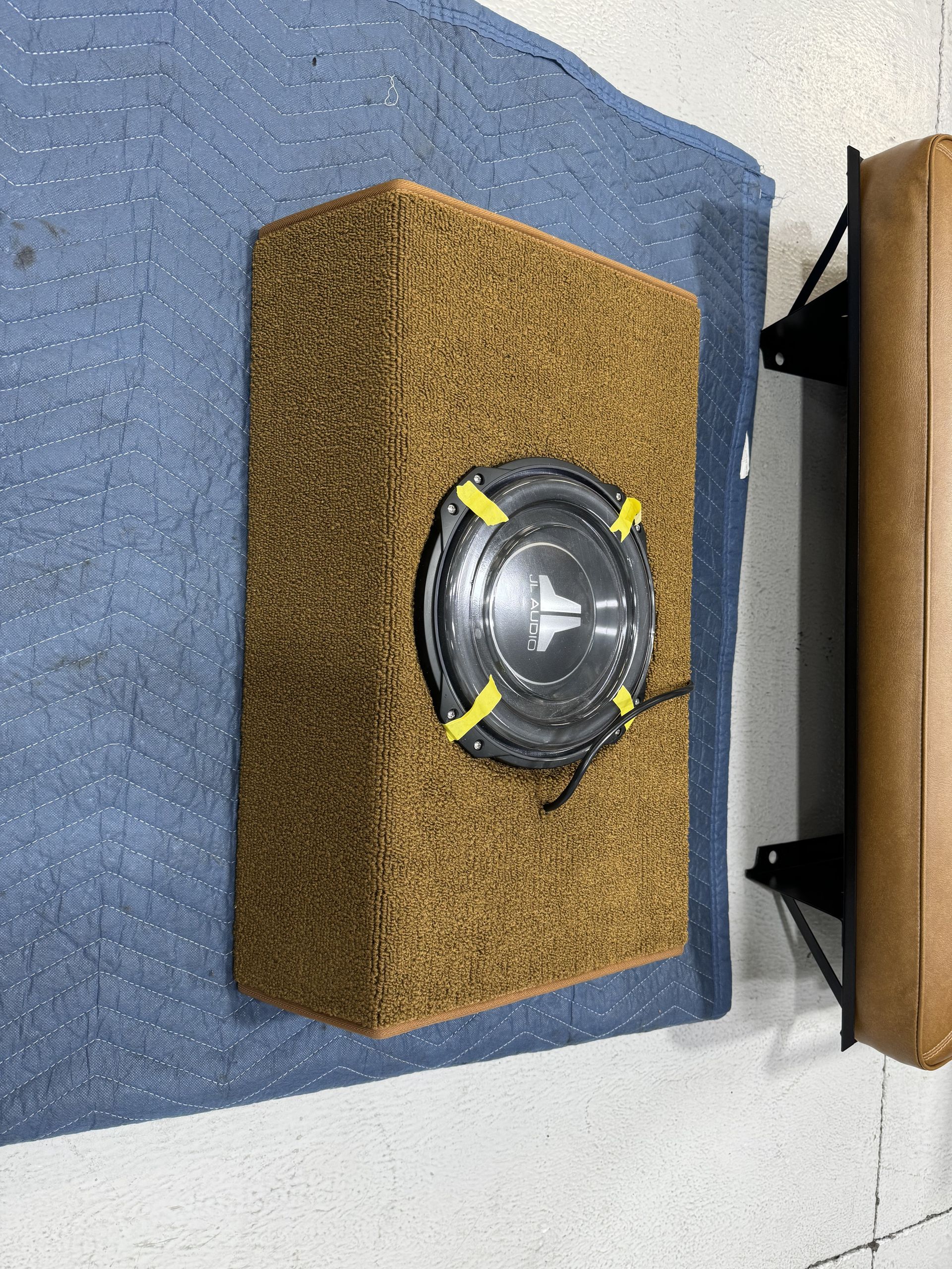 Speaker in a small wooden box on a blue carpeted floor, viewed from above.