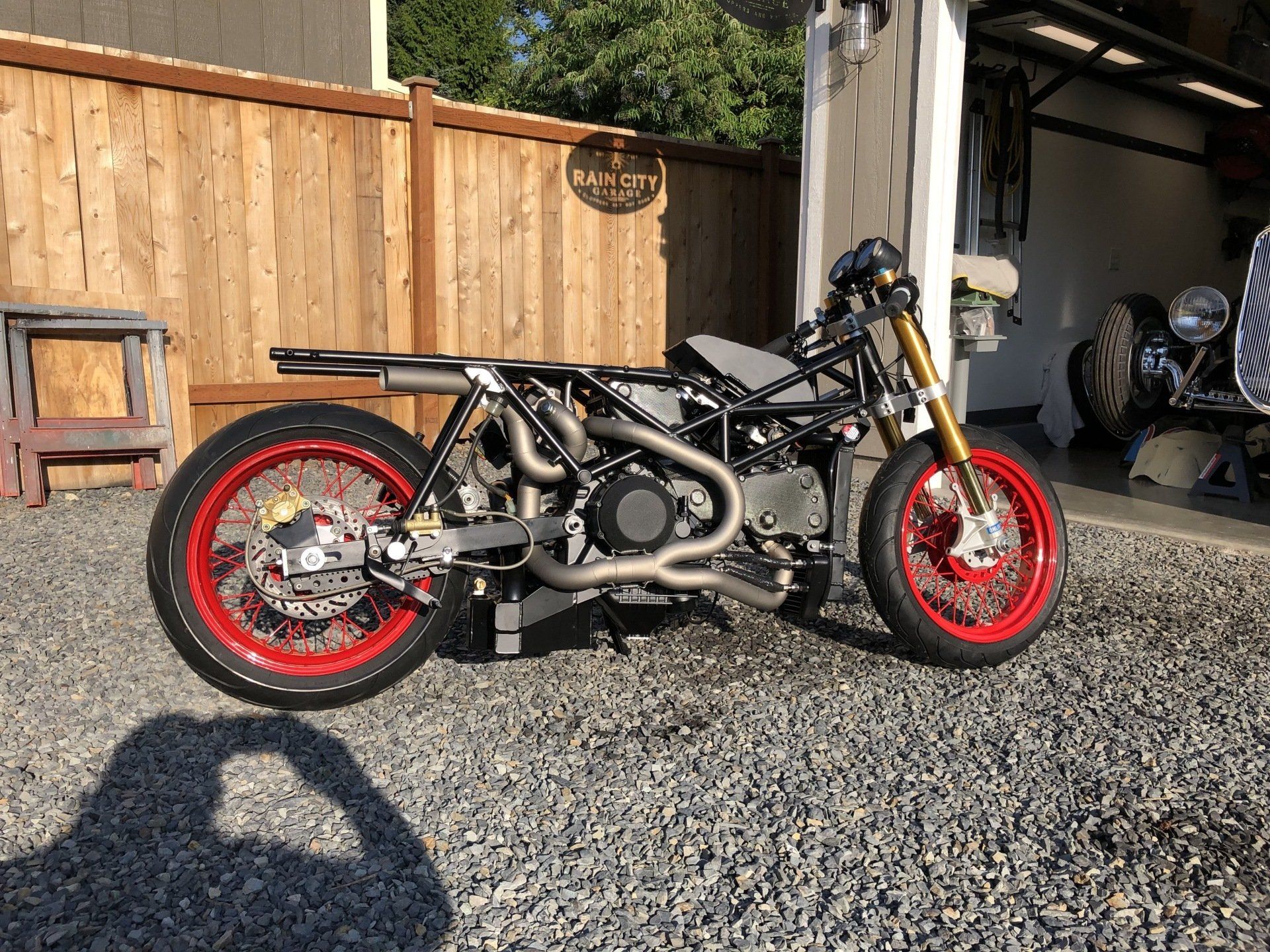 Custom black motorcycle with red wheels parked on gravel beside a wooden fence and garage.