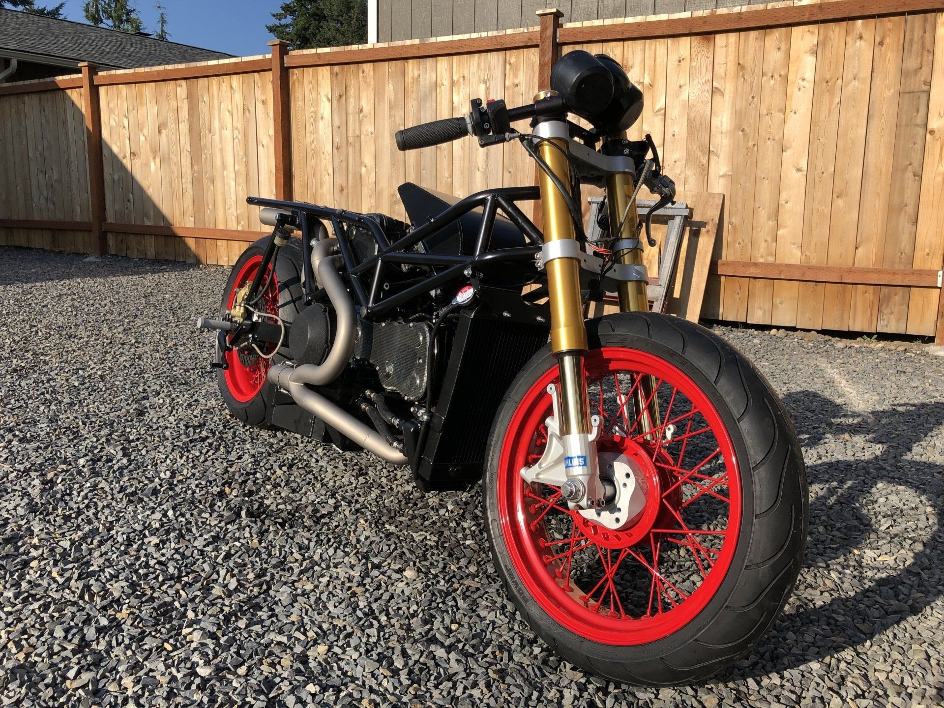 Custom black motorcycle with red wheels parked on gravel beside a wooden fence.